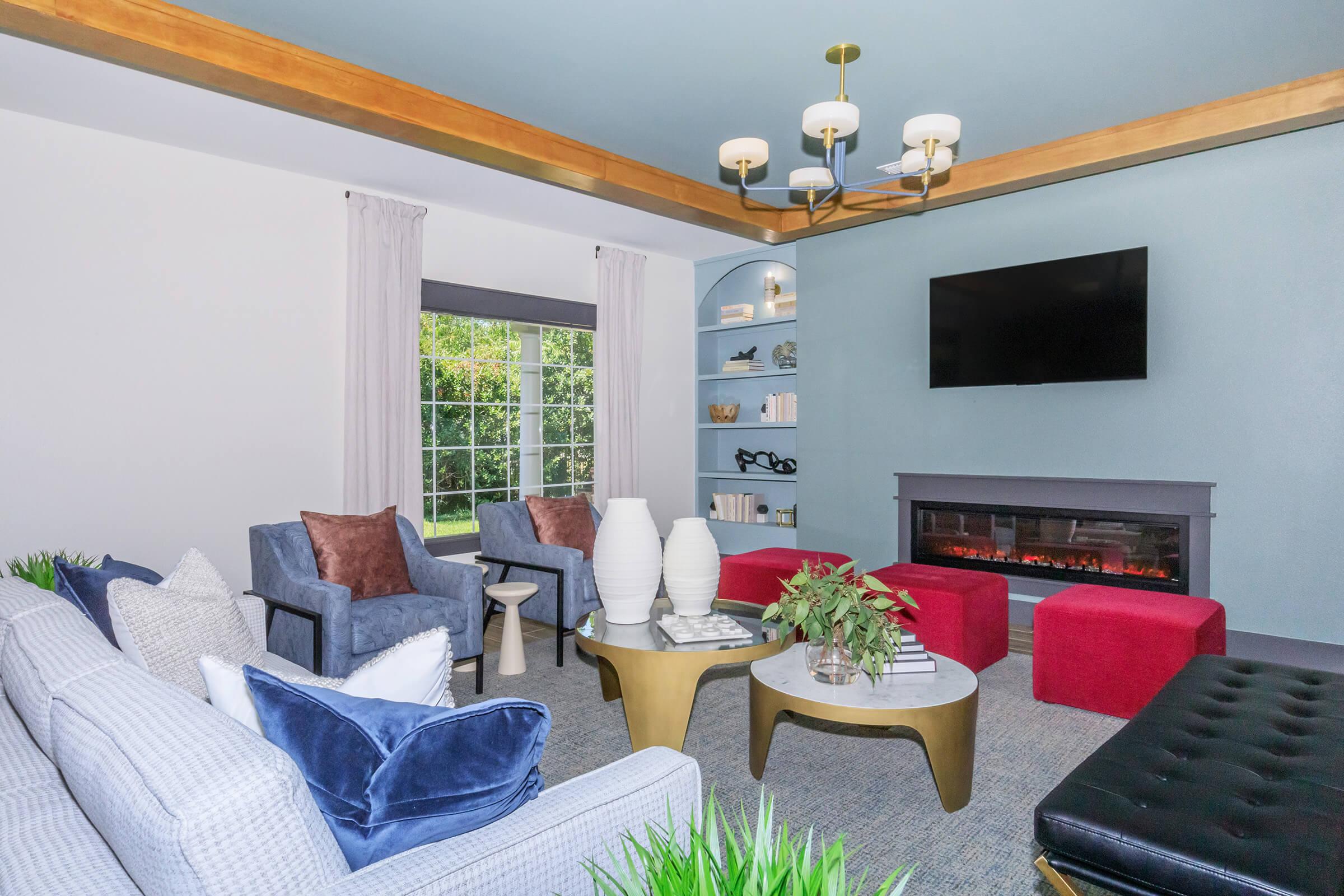 A stylish living room featuring a gray sofa with blue and white pillows, two blue armchairs, a modern coffee table, and an electric fireplace. Large windows allow natural light, creating a cozy atmosphere. Decorative elements include potted plants and a chandelier above.