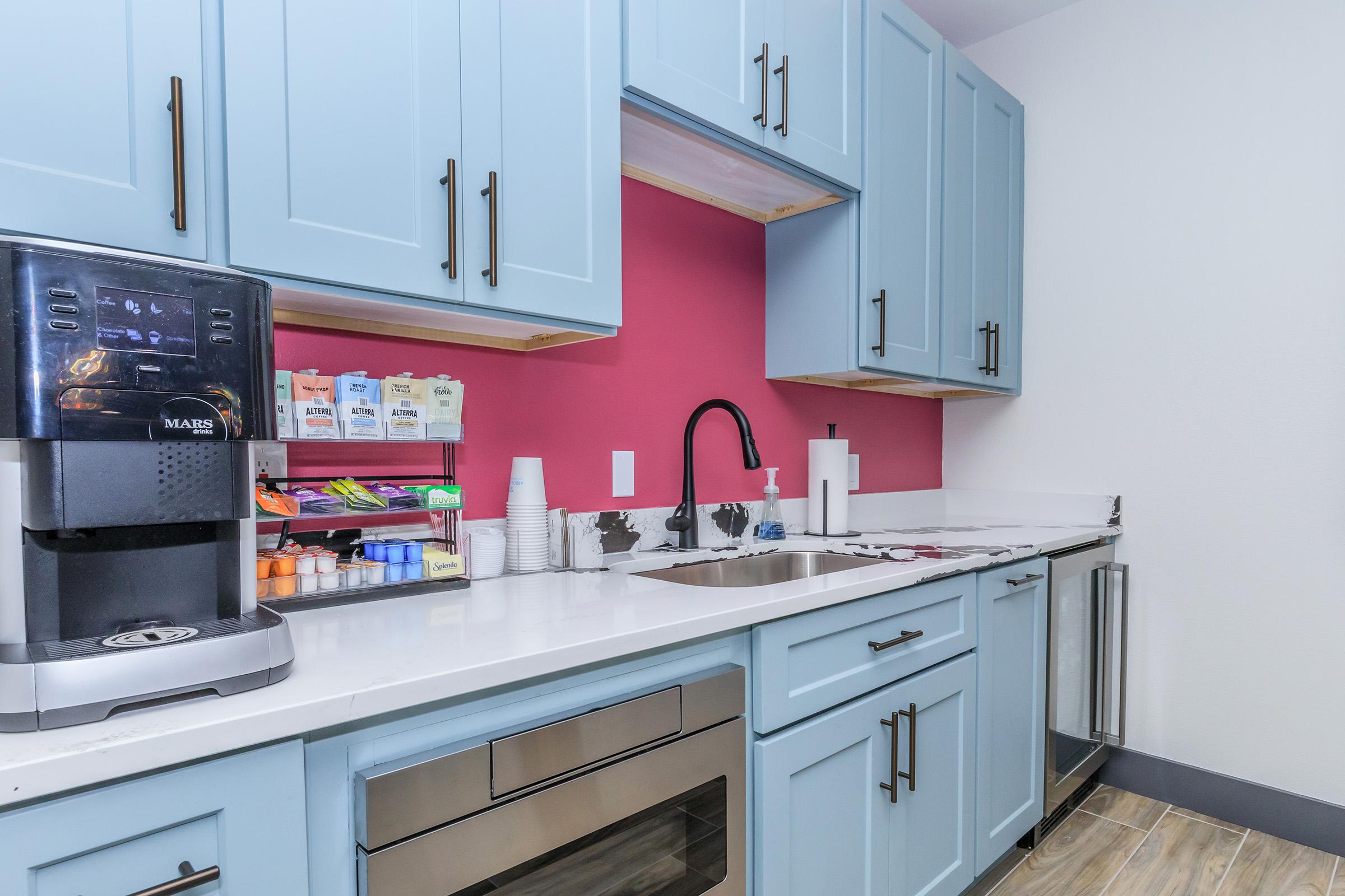 A modern kitchen featuring light blue cabinets and a vibrant pink wall. There is a coffee machine on the counter, alongside an organized assortment of coffee supplies and tea bags. A sleek sink and a stainless steel refrigerator are visible, enhancing the contemporary design of the space.