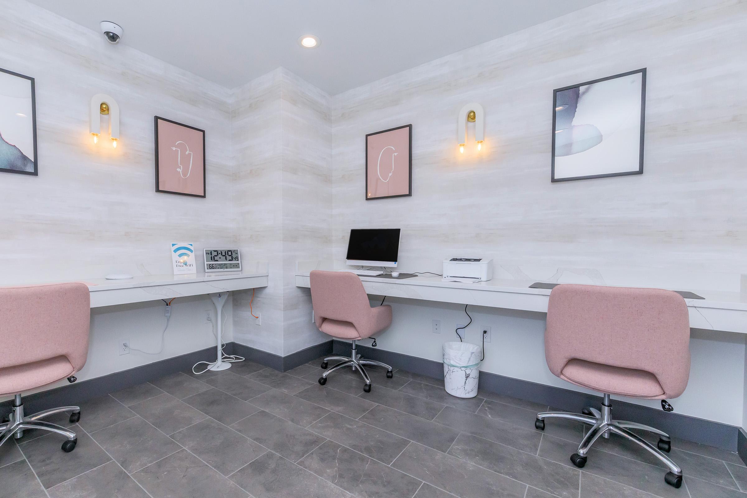 Two modern workspaces in a well-lit room featuring light-colored walls and soft pink chairs. Each station is equipped with a desk and computer. Wall art in minimalistic frames adds a touch of decor. The space also contains a wall clock and a trash bin, enhancing its functional and aesthetic appeal.