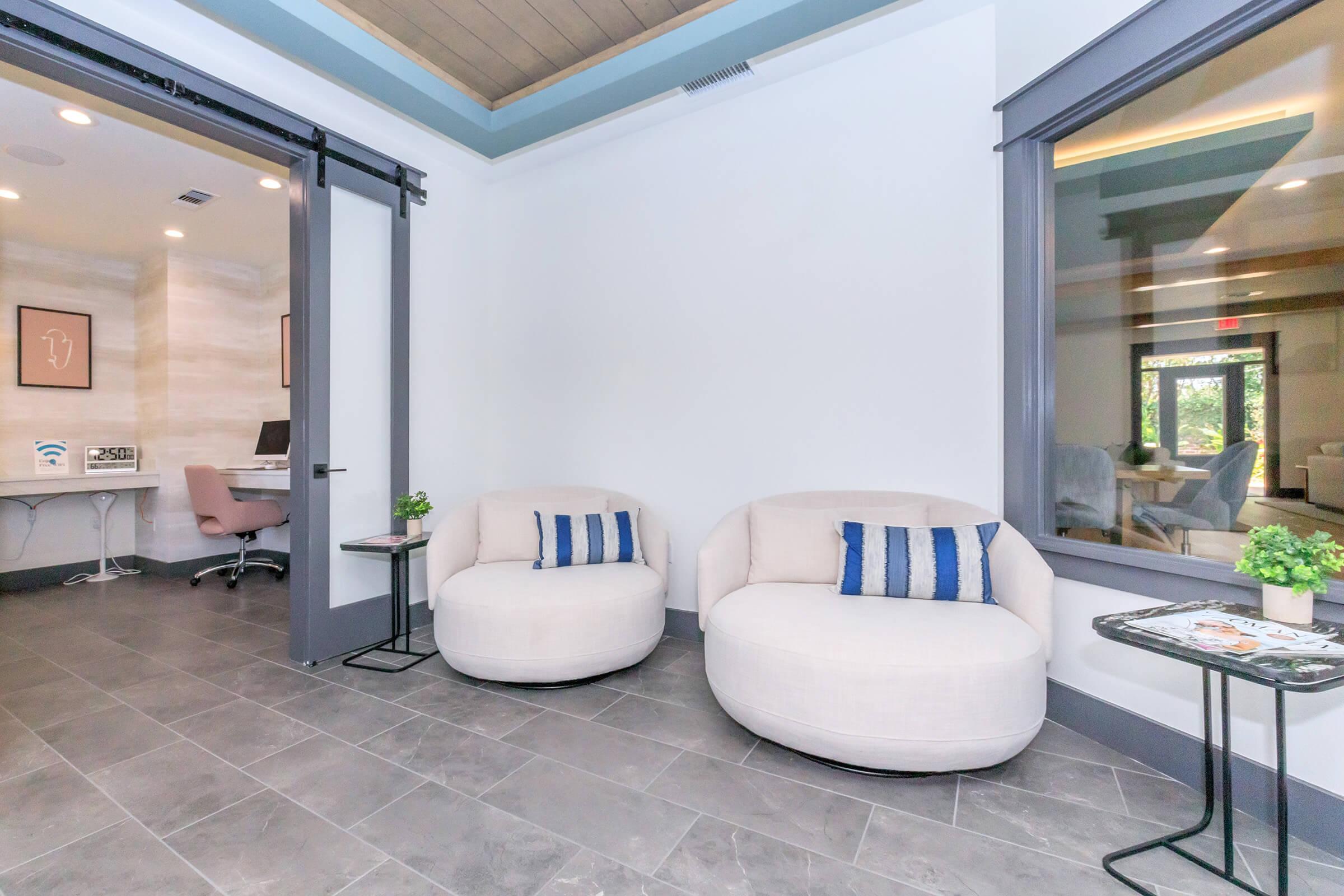 A modern lounge area featuring two round, beige armchairs with blue-striped pillows. Small tables with plants are beside the chairs. In the background, a glass-walled office space is visible, with a desk and chair. The flooring is gray tile, and the ceiling has wooden panels.