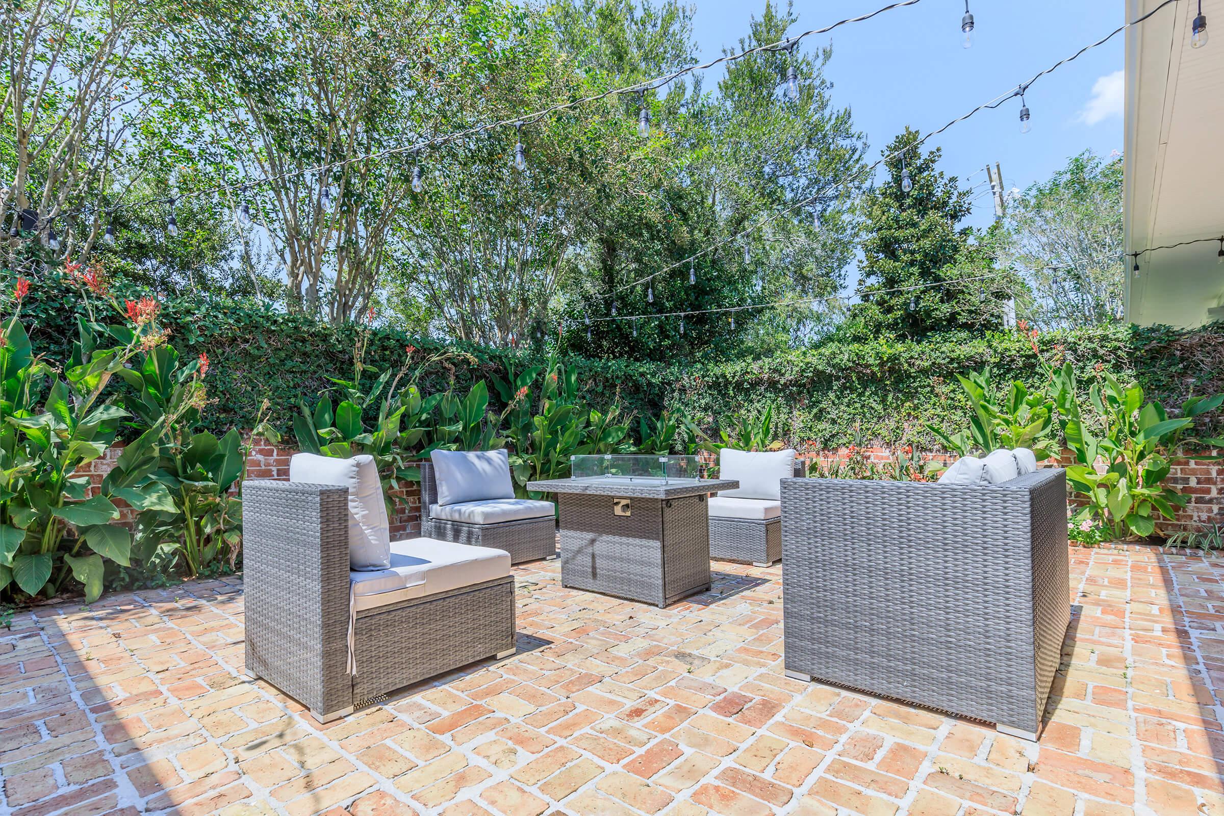 A cozy outdoor seating area featuring four woven grey chairs with light cushions around a square glass-top table. The space is surrounded by lush greenery and flowering plants, set against a backdrop of a brick pathway, under a clear blue sky with string lights overhead.