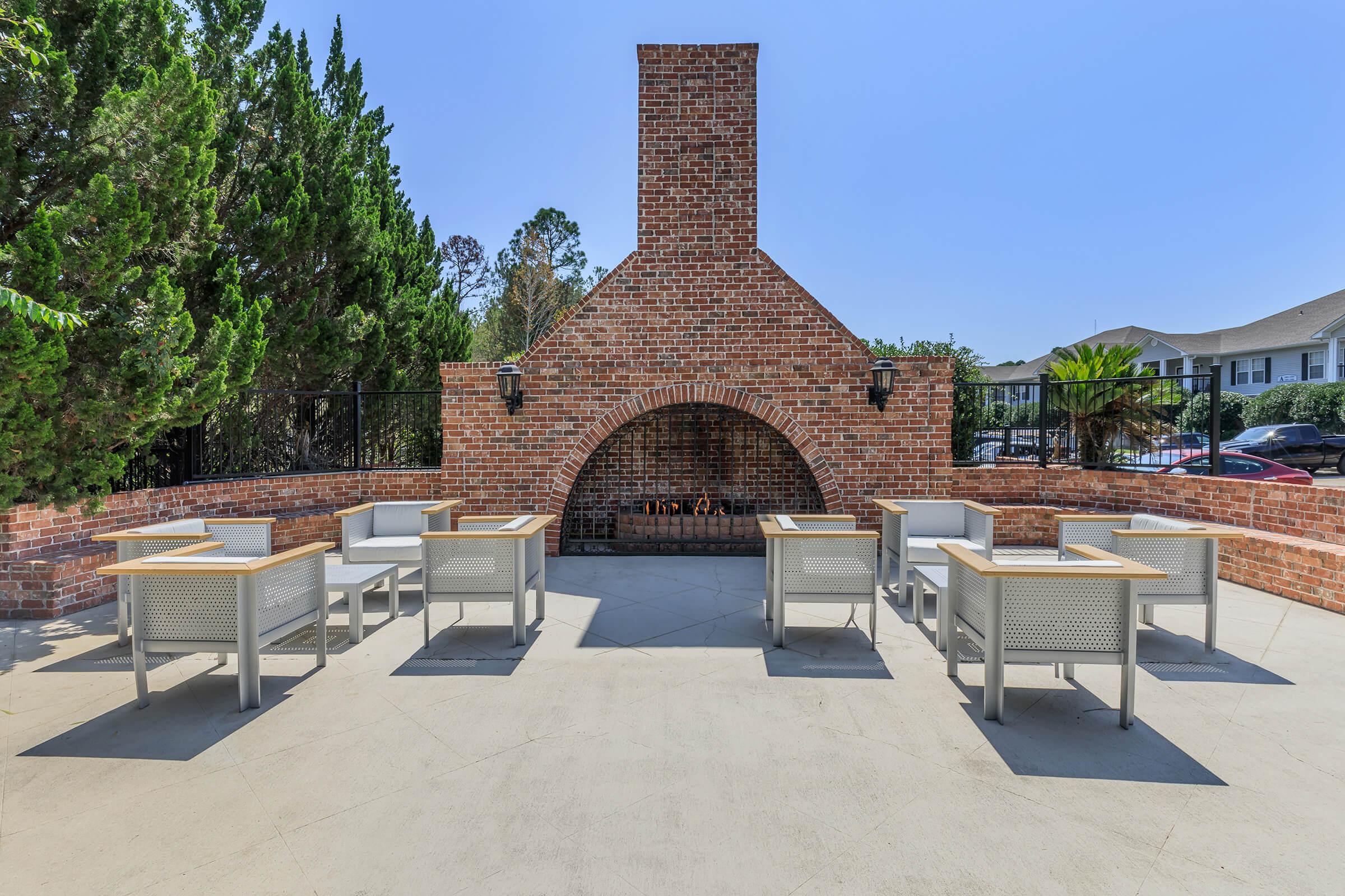 Outdoor patio featuring a large brick fireplace, surrounded by several tables with modern chairs. The area is lined with trees, and there is a clear blue sky overhead, creating a welcoming space for gatherings.