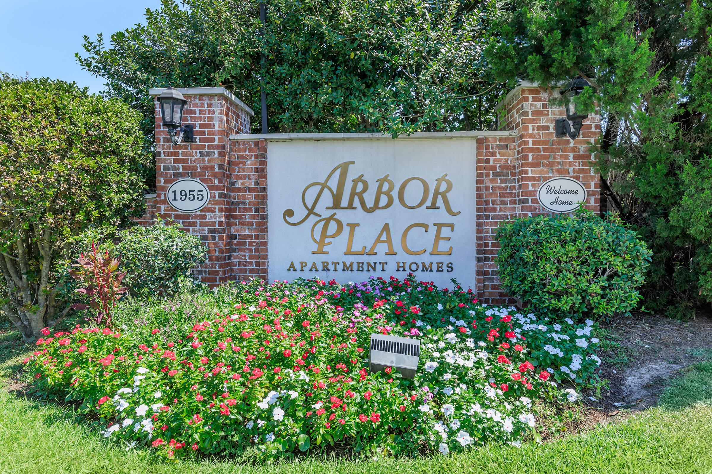 Sign for Arbor Place Apartment Homes, featuring a brick structure with lush green shrubs and colorful flower beds. The sign displays "Arbor Place" prominently, with "Apartment Homes" underneath, and the year "1955" on one side. The overall setting is welcoming and well-maintained.