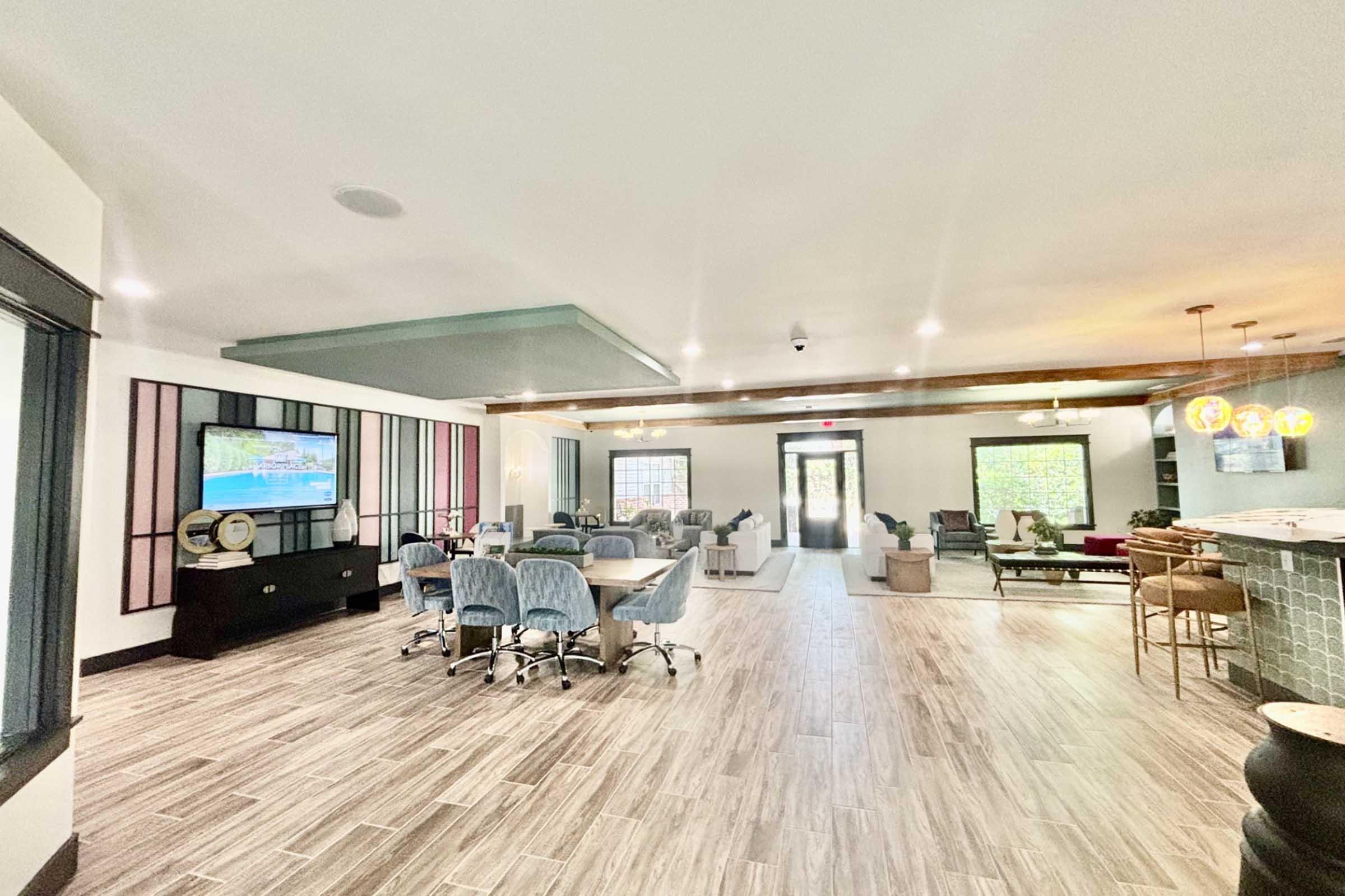 A spacious interior featuring a modern lounge area with various seating options, a large TV on the wall, and a decorative counter. Natural light flows through large windows, and potted plants add a touch of greenery. The flooring is designed with wood-like tiles, creating a warm and inviting atmosphere.