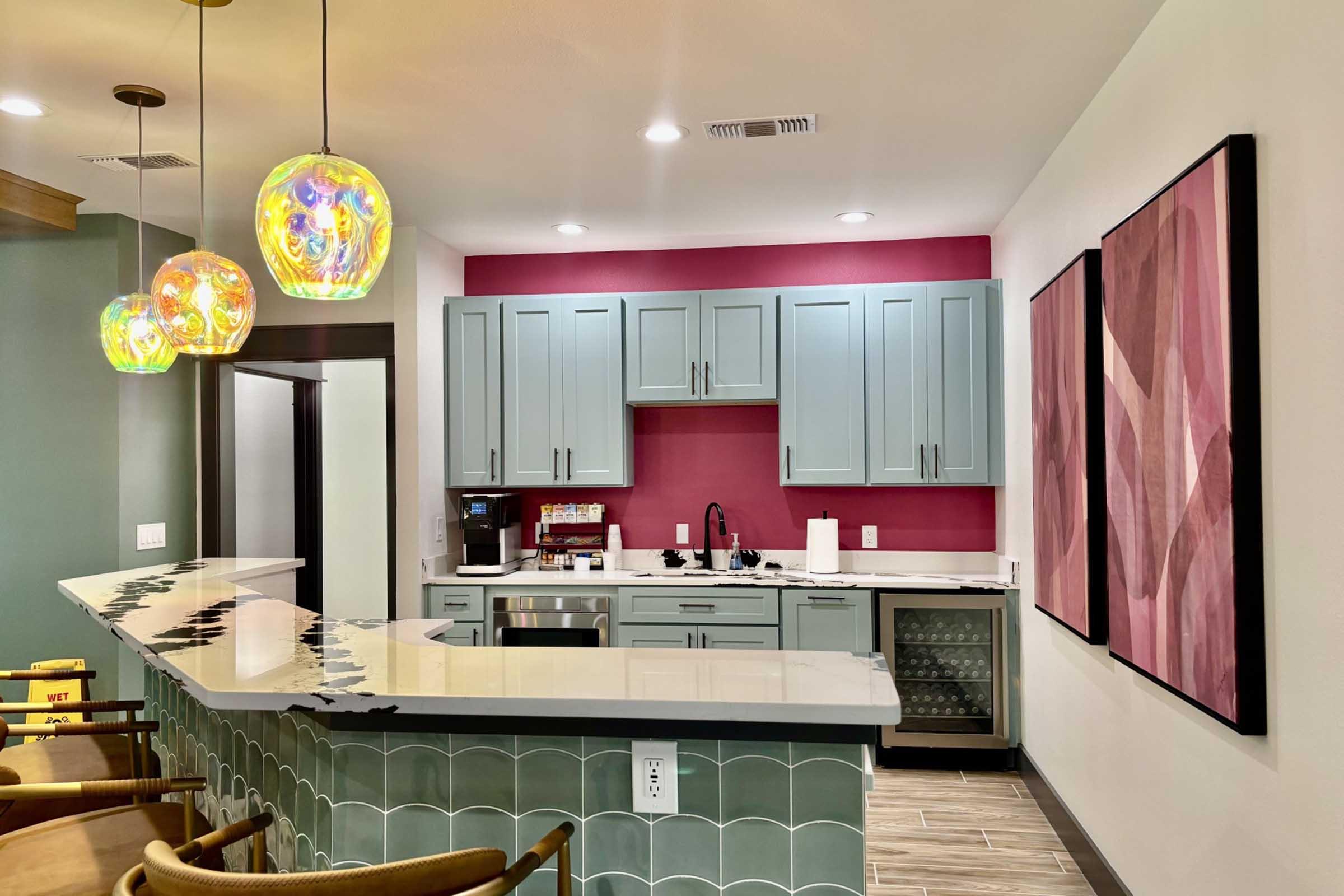 Modern kitchen with light blue cabinets and a pink accent wall. Features a marble countertop with a white and gray design, a decorative tiled front on the bar, and colorful pendant lights. Includes a coffee maker, sink, and a small wine fridge. Two abstract art pieces adorn the wall.
