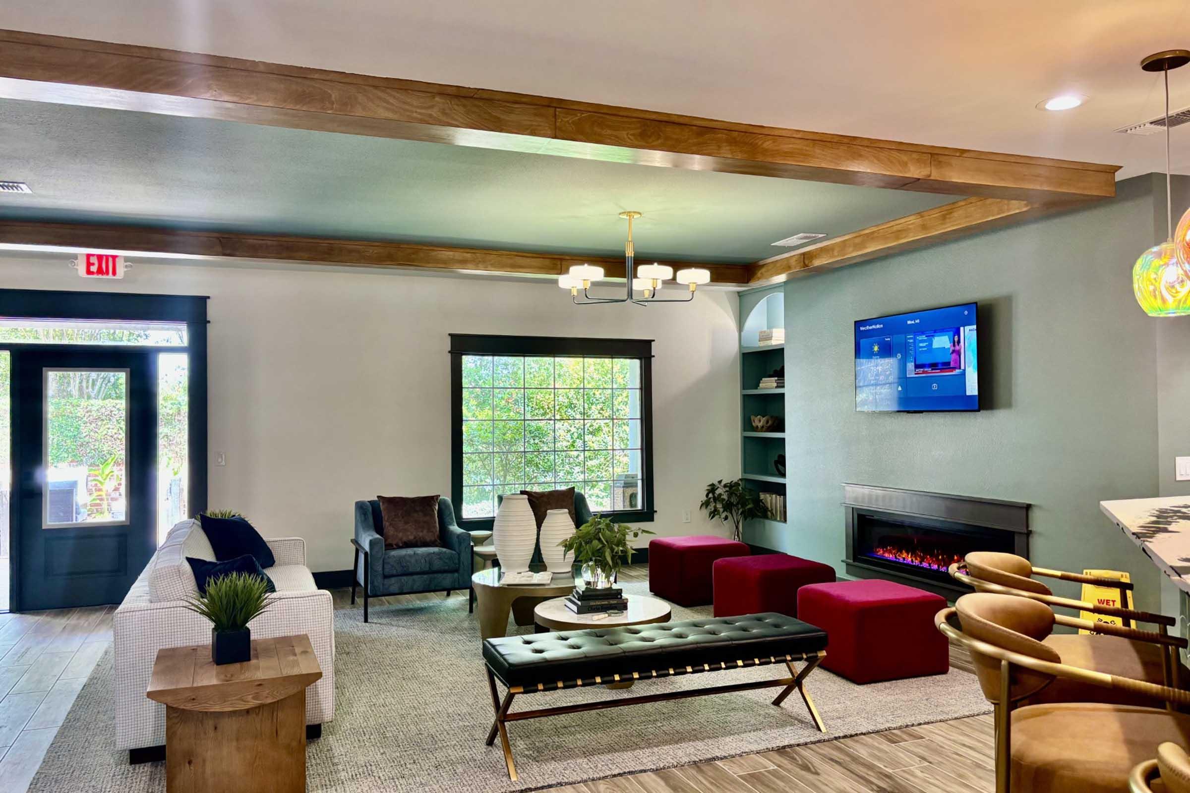 A modern living room featuring a cozy seating area with a white couch, dark blue armchair, and a black ottoman. There's a fireplace, a large window with natural light, and a flat-screen TV on the wall. Stylish decor includes a potted plant and a round coffee table, creating an inviting atmosphere.