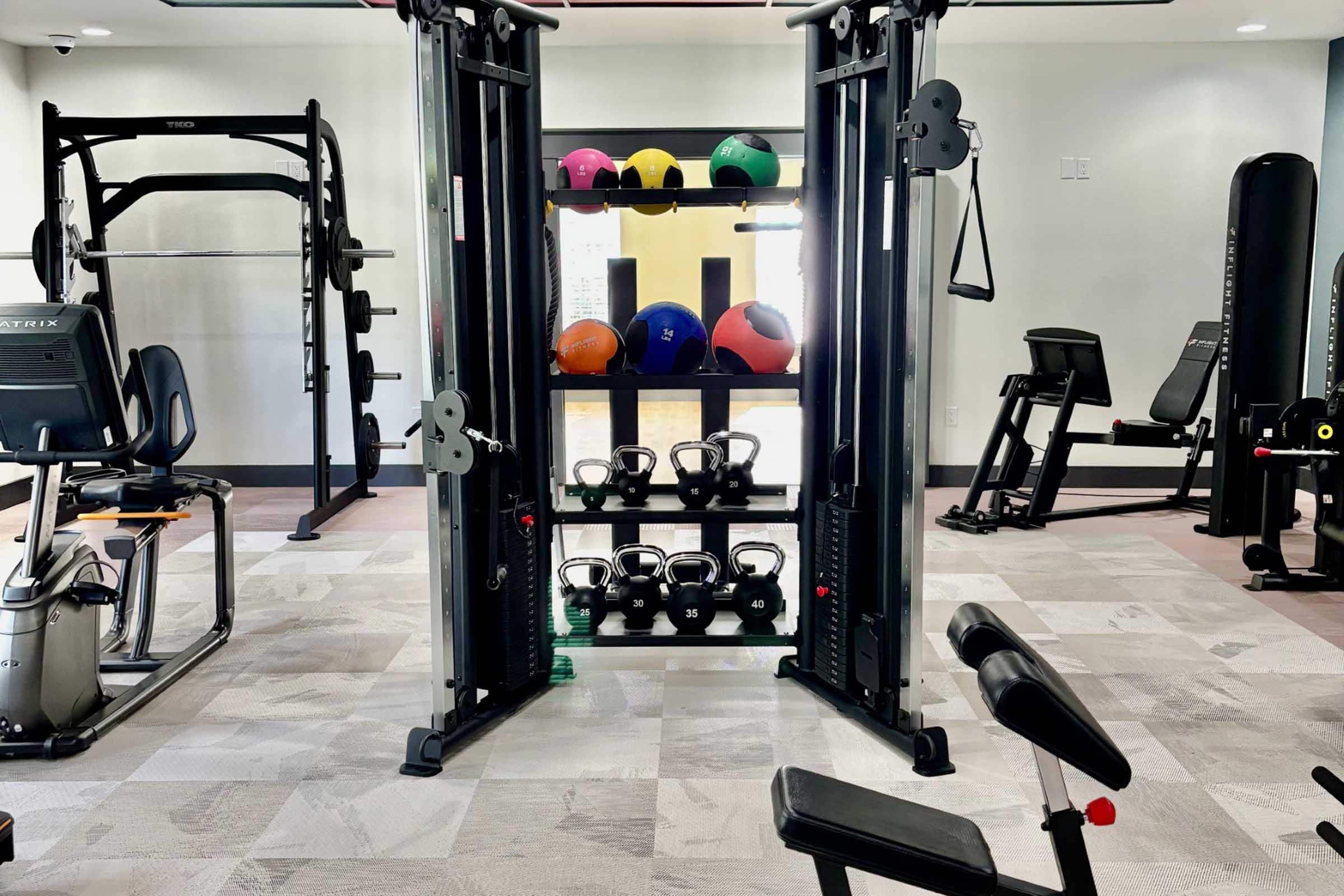 A modern gym interior featuring a cable machine in the center, surrounded by various gym equipment, including free weights, kettlebells, and exercise balls stacked on a shelf. The floor is covered with light-colored tiles, creating a spacious and inviting workout space.