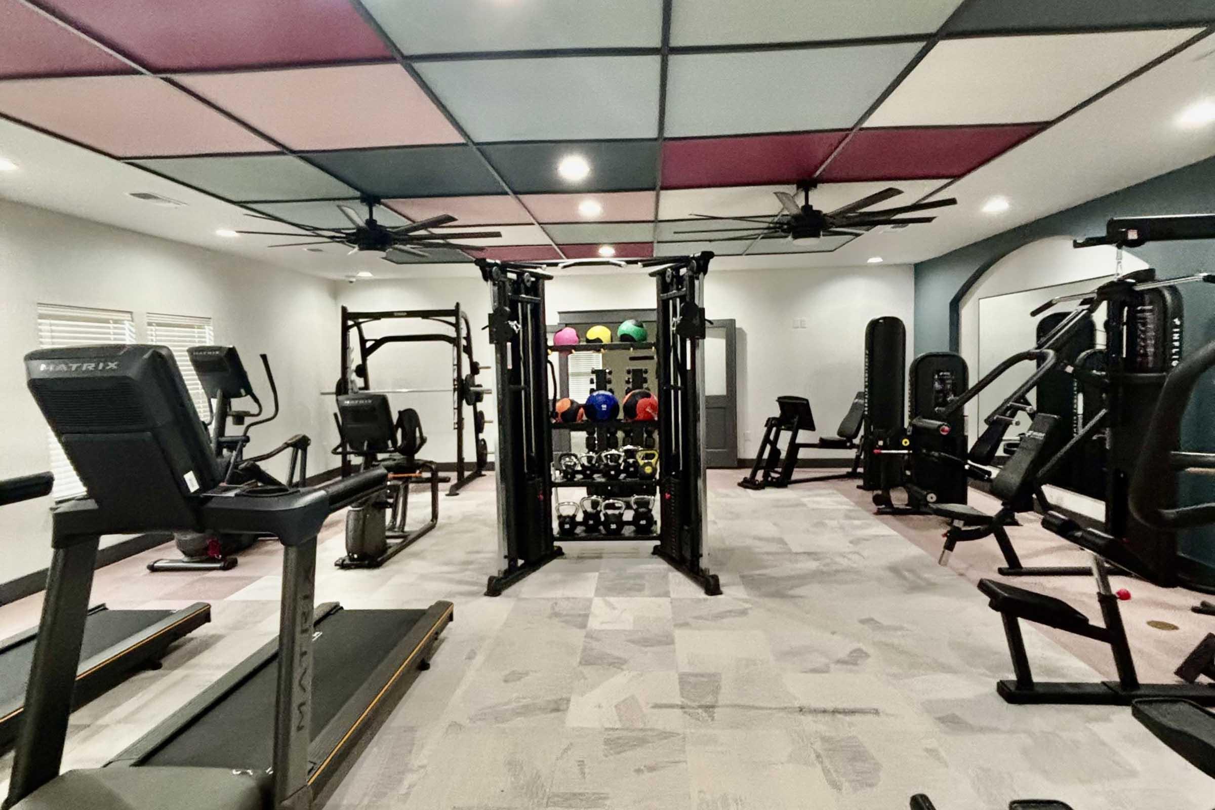 A well-equipped home gym featuring various fitness machines, including treadmills, a multi-gym weight station, and a rack of colorful dumbbells. The room has a modern design with a unique ceiling and adequate lighting, creating an inviting workout environment.