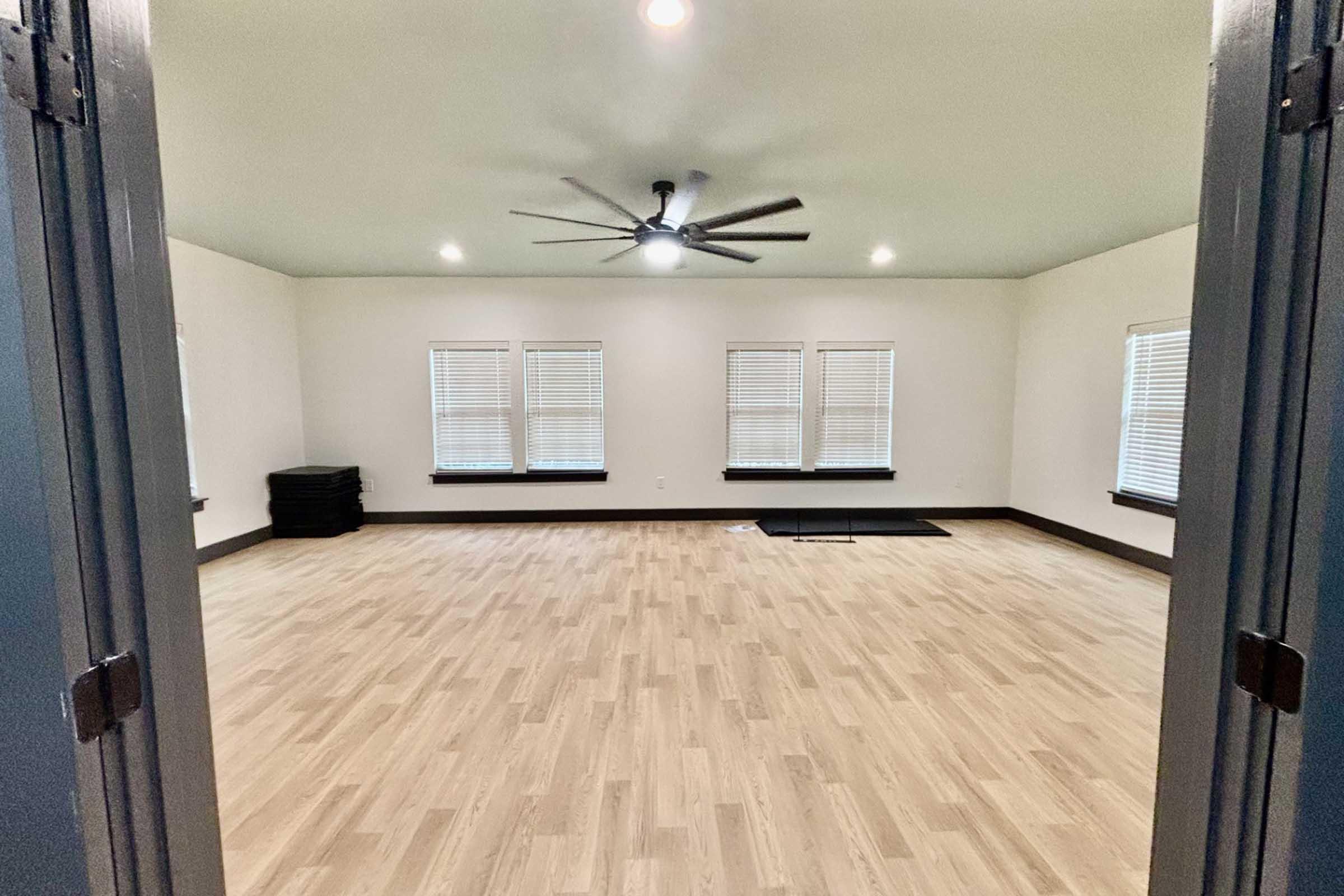 A spacious, well-lit room with light wood flooring and white walls. It features three windows with blinds, a ceiling fan, and a few black mats stacked in the corner, creating an open and inviting atmosphere suitable for activities or gatherings.
