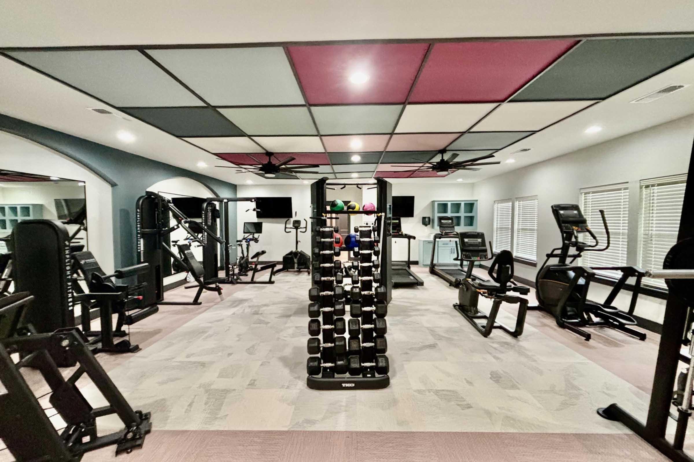 A spacious home gym featuring various exercise equipment including weightlifting stations, dumbbells, a treadmill, and an elliptical machine. The room has a modern design with a stylish ceiling and good lighting, providing an inviting space for workouts.