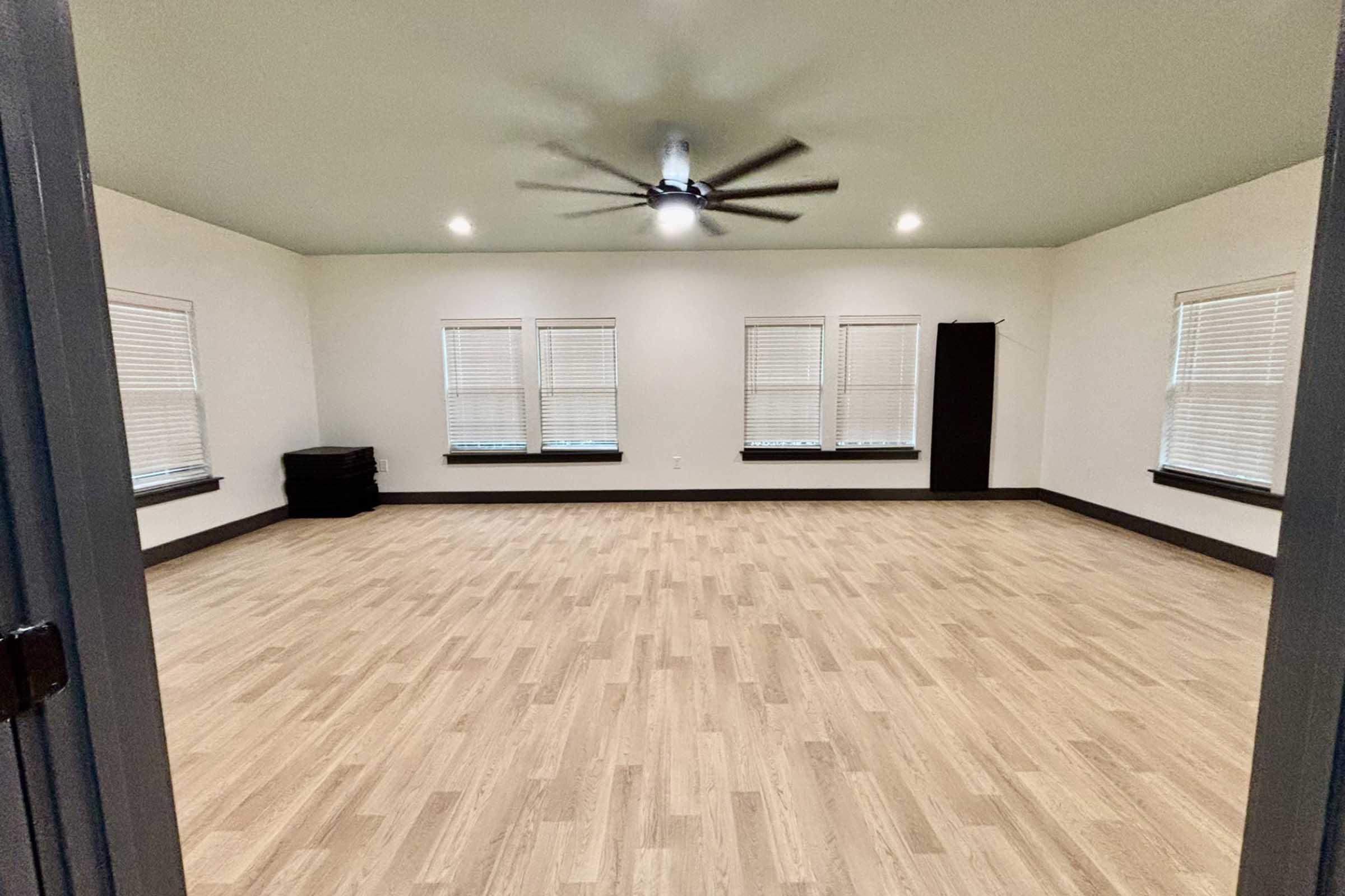 A spacious empty room with light-colored wooden flooring, several windows with blinds allowing natural light, and a ceiling fan. The walls are painted light, and a dark storage unit is visible in one corner. The layout suggests versatility for various uses, such as a meeting space or studio.