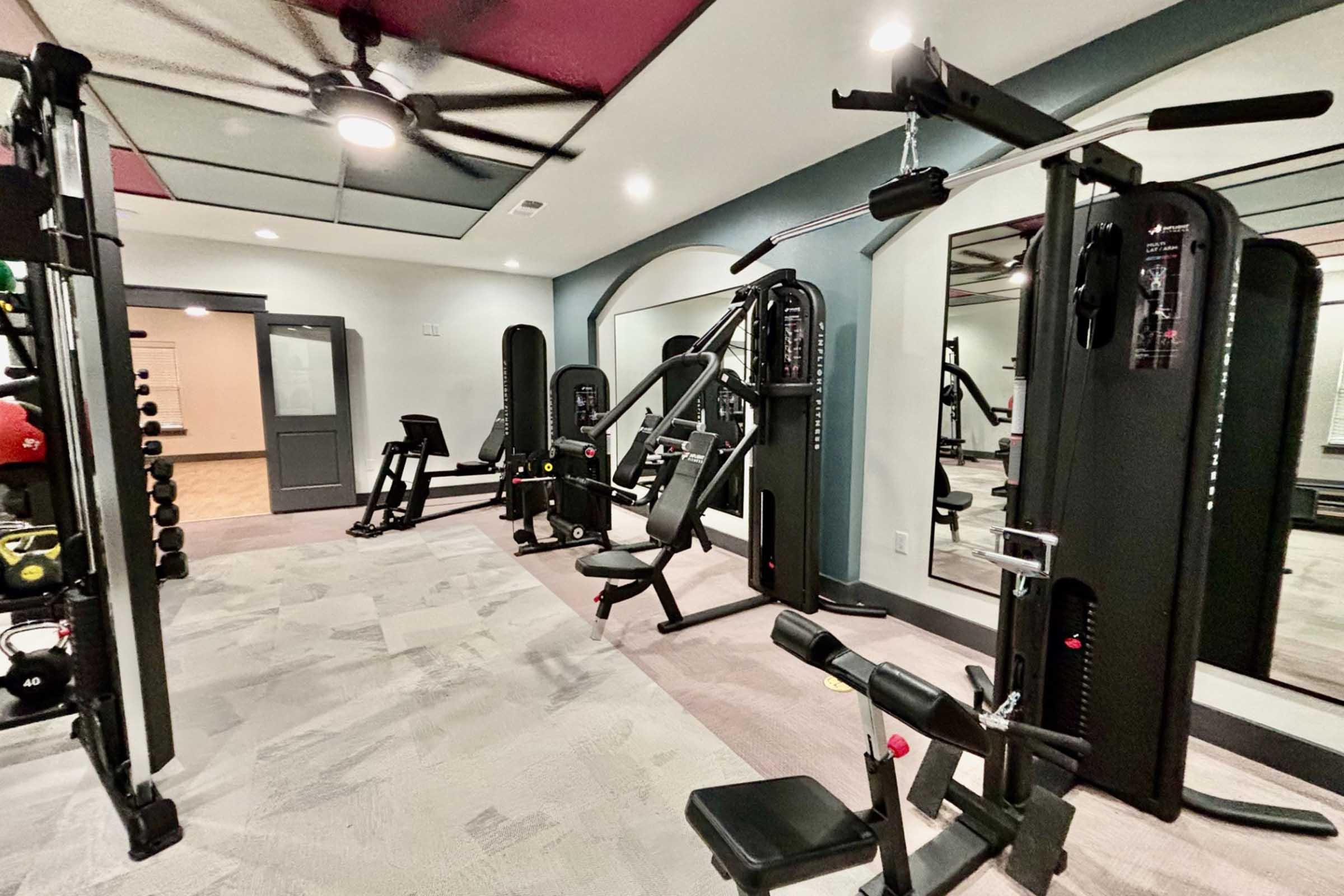 A modern gym featuring various workout equipment, including multi-station strength machines, a bench press, free weights, and a large mirror on the wall. The space is well-lit, with a ceiling fan and a neutral-colored floor, creating an inviting environment for exercise.