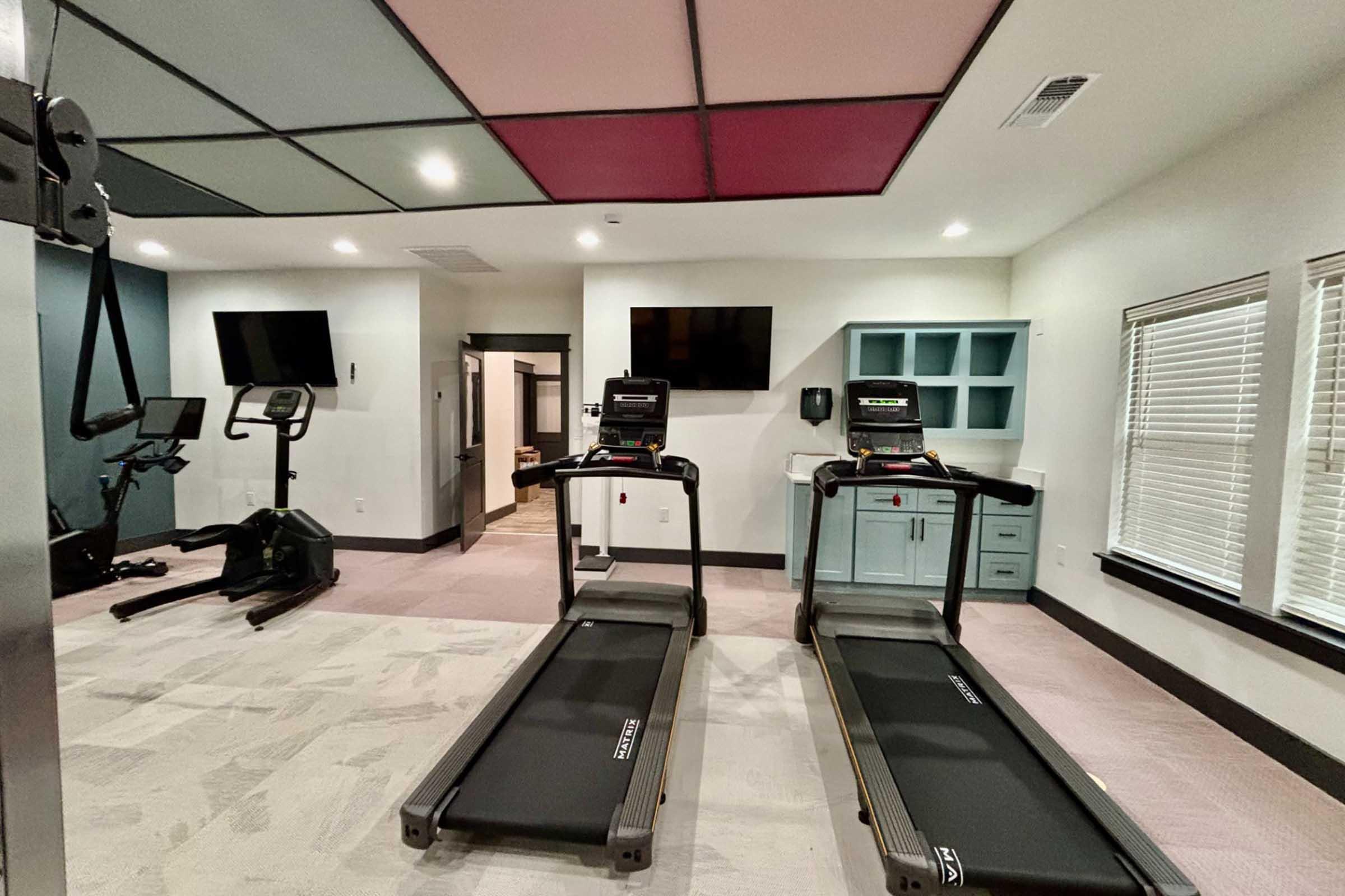 A modern fitness room featuring two treadmills, an exercise bike, and two wall-mounted televisions. The ceiling is divided into colorful panels, and there is a turquoise cabinet along one wall. Bright lighting enhances the space, which has large windows allowing natural light to enter.