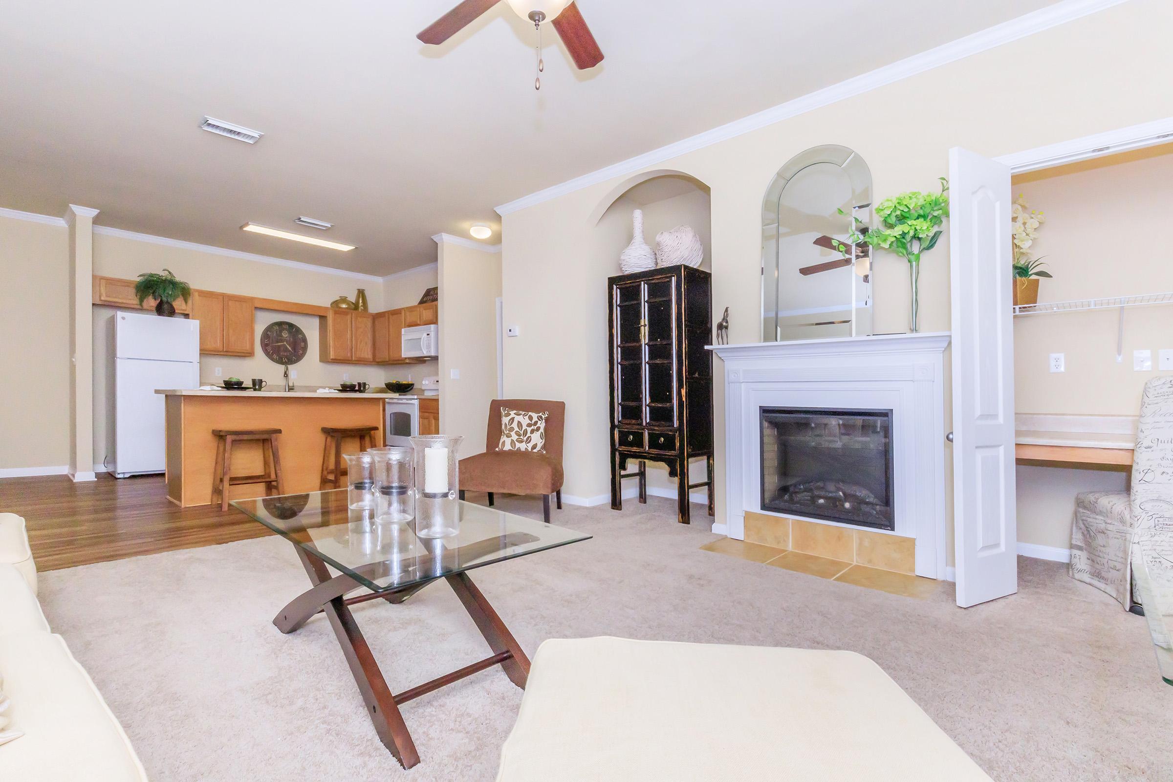 A cozy living room with a light beige color scheme, featuring a glass coffee table, a comfortable sofa, a ceiling fan, and a small fireplace. The room opens to a kitchen area with wooden cabinets and a refrigerator. A decorative cabinet and plants add a welcoming touch.