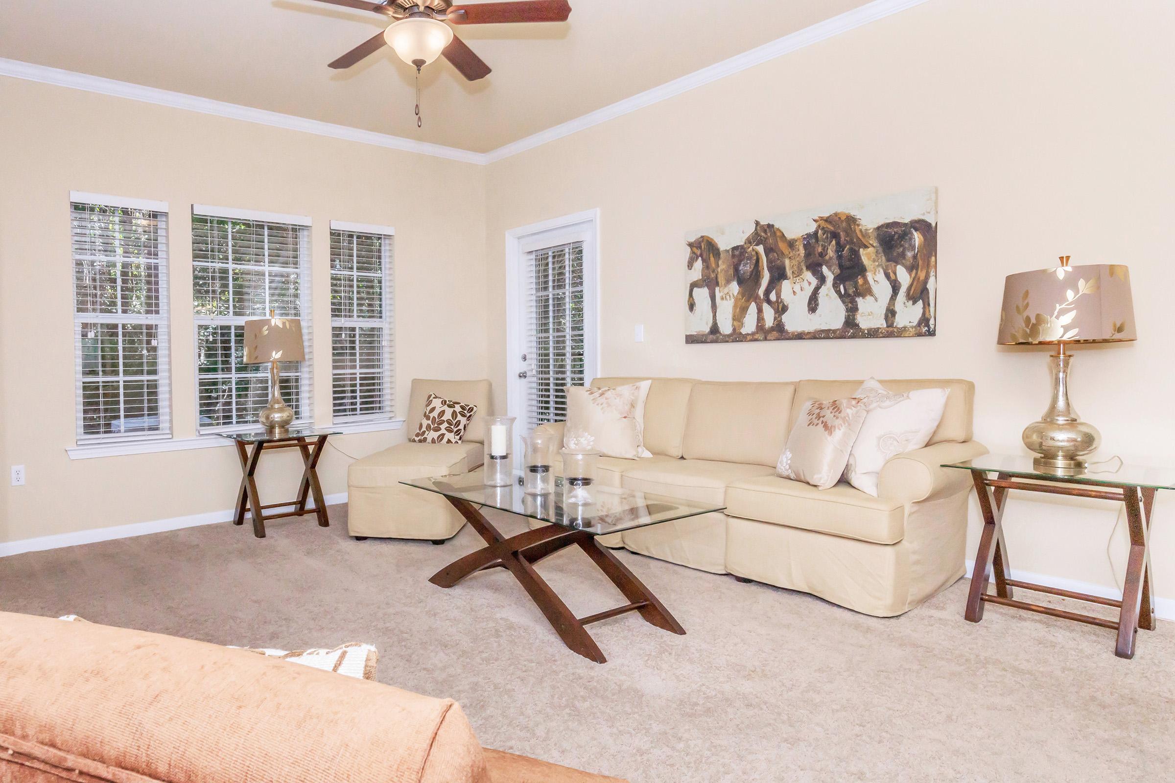 A cozy living room featuring a beige sofa with decorative pillows, two glass coffee tables, and a pair of elegant lamps on wooden side tables. Natural light streams in through multiple windows. A painting of horses adorns the wall, adding a touch of art to the serene atmosphere.