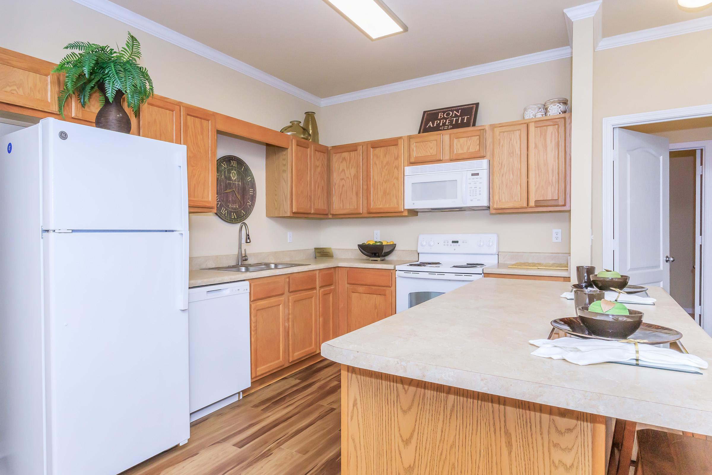 Modern kitchen featuring wooden cabinets, a white refrigerator, and a microwave. Countertops are beige with decorative bowls and plants. A dining setting is arranged with plates and utensils on the island. The wall has a "Bon Appétit" sign, and there's a clock for added decor. Bright and inviting atmosphere.