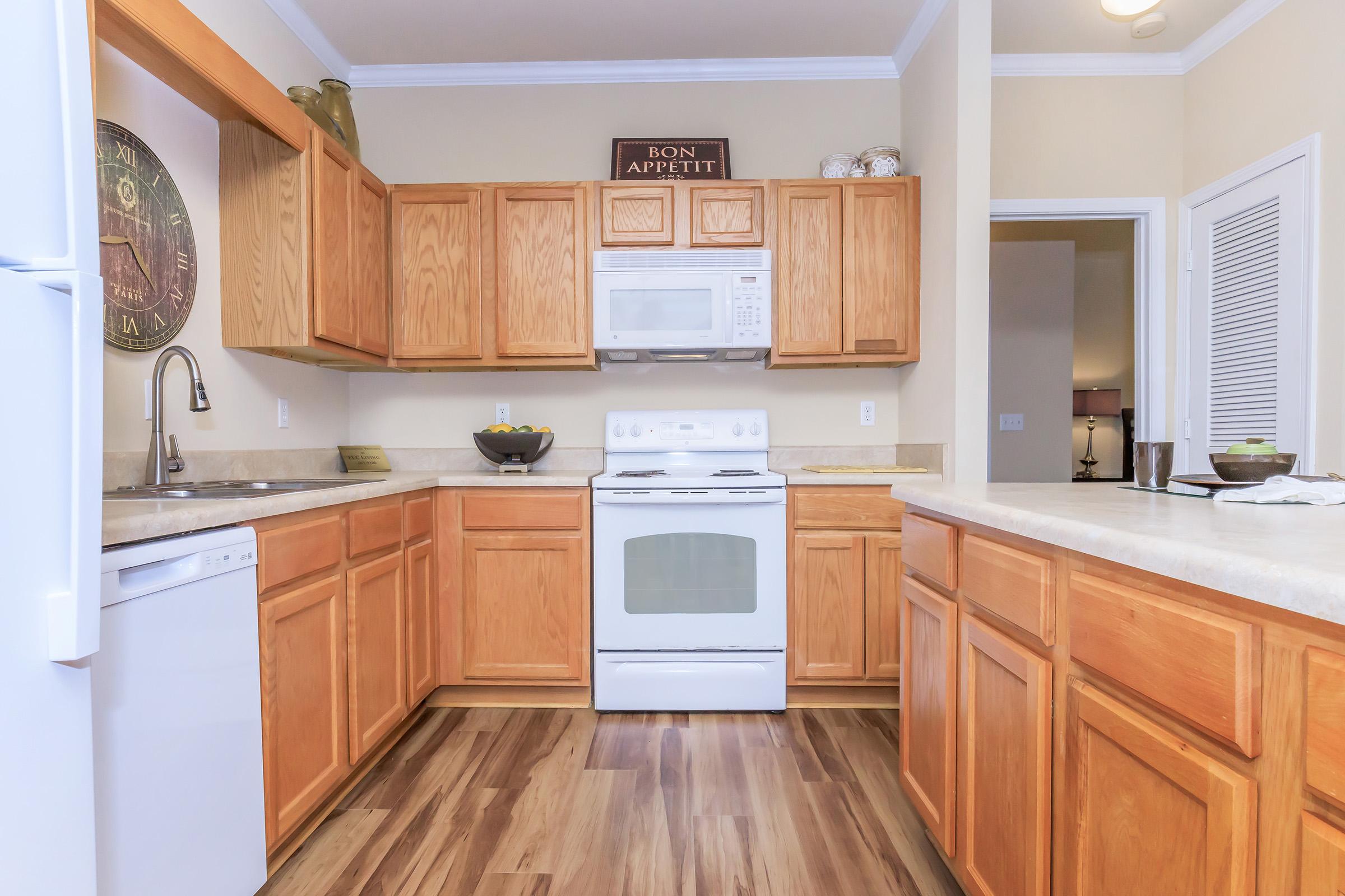 A modern kitchen featuring wooden cabinets and a beige countertop. Includes a white appliance suite with a microwave, oven, and dishwasher. A wall-mounted clock and decorative items add character, and the floor has a warm wood-like finish. Bright, inviting atmosphere with neutral-colored walls.