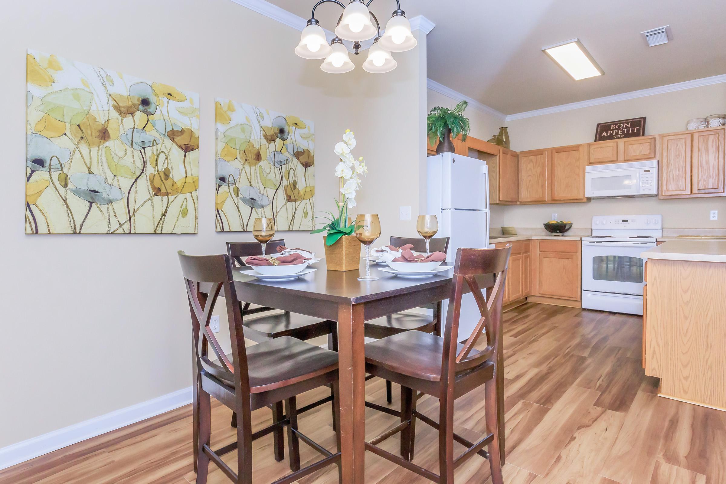 A cozy modern kitchen with a small dining table set for two. The table features decorative plates and glasses. The walls have floral artwork, and there’s a light fixture above. The kitchen area includes wooden cabinetry, a stove, and a refrigerator, creating a warm and inviting atmosphere.