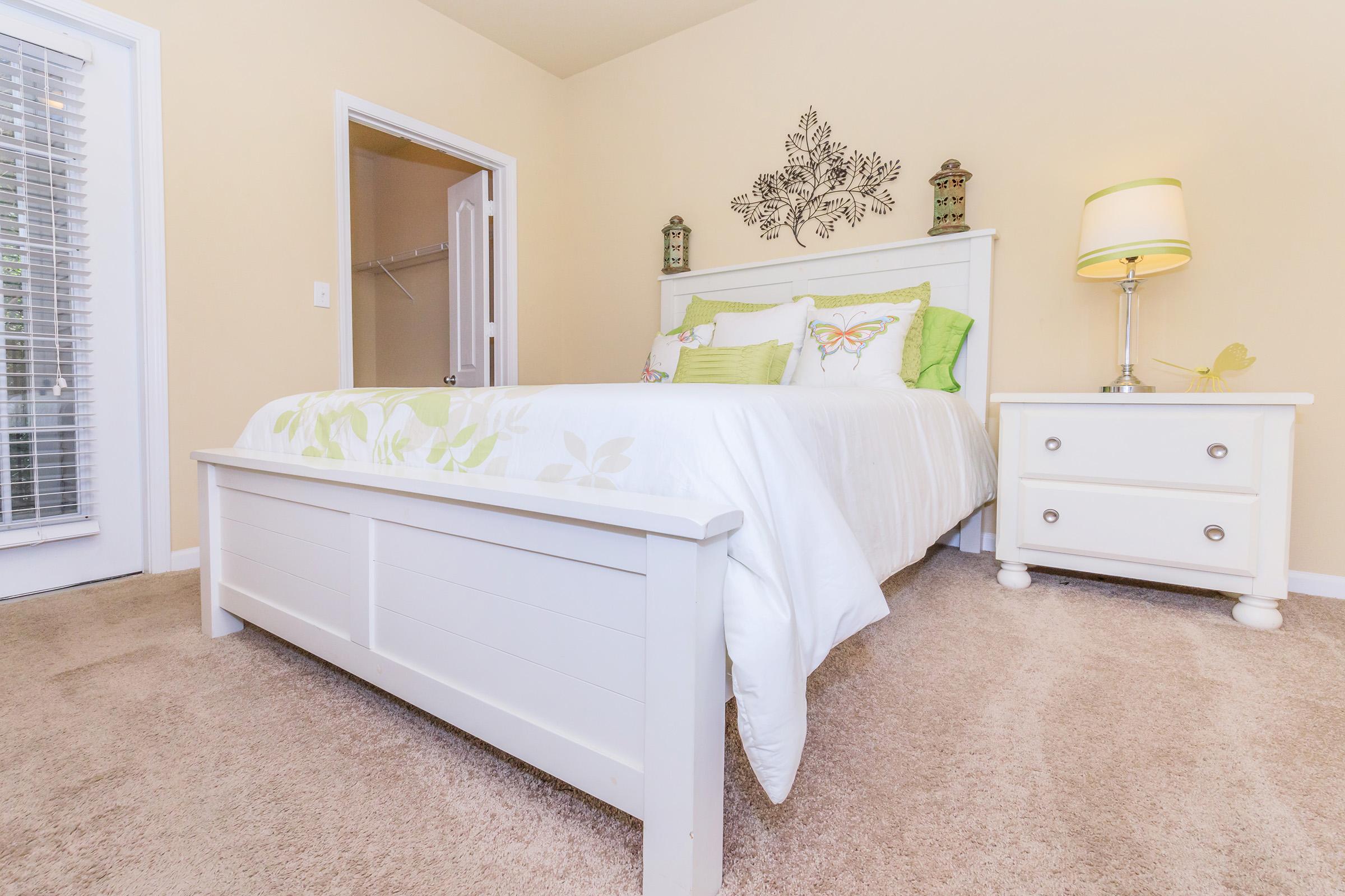 A bright bedroom featuring a white bed with decorative pillows, a bedside table with a lamp, and light-colored walls. There is a closet door visible, and the floor is carpeted in a soft, neutral shade. A decorative wall piece adds a touch of elegance to the space.