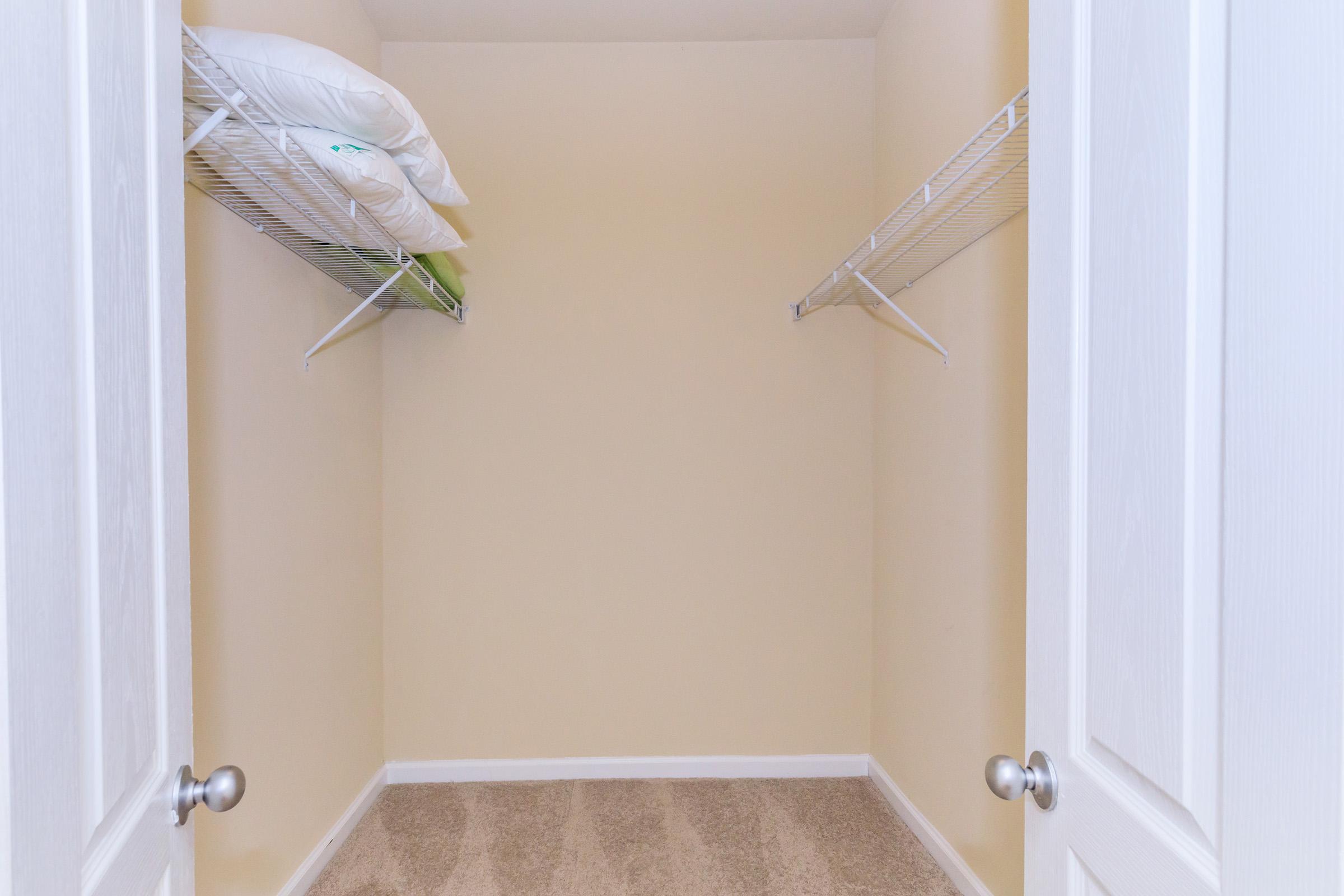 A clean, empty closet with two wire shelves. One shelf has two pillows neatly placed on it, while the other shelf is vacant. The walls are painted a light color, and the floor is carpeted. The closet doors are open, allowing full view of the interior.