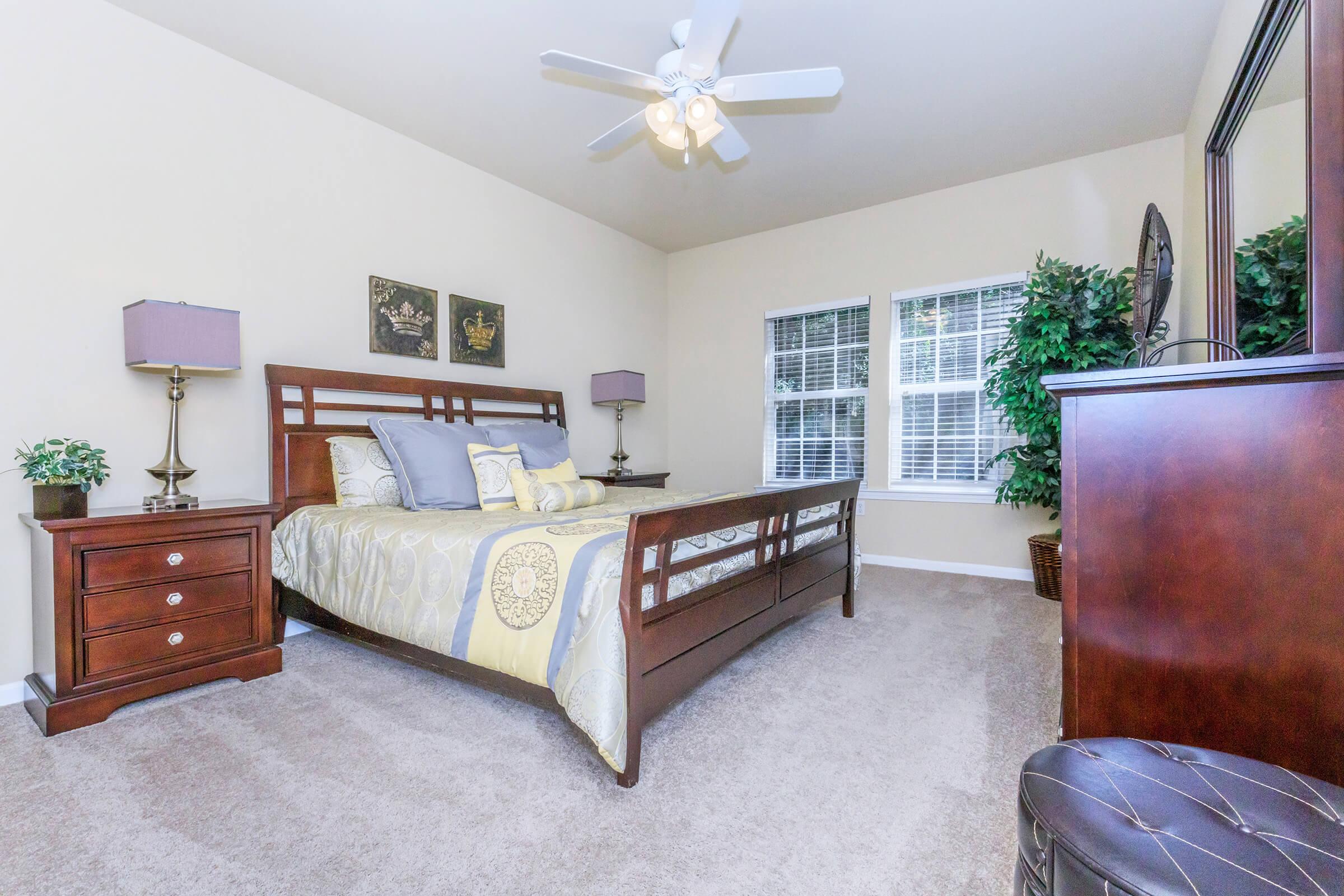 A well-decorated bedroom featuring a wooden bed with decorative bedding, two bedside lamps on matching nightstands, windows with blinds, and a large dresser. The room has plush beige carpeting and a decorative plant in the corner, creating a cozy atmosphere.