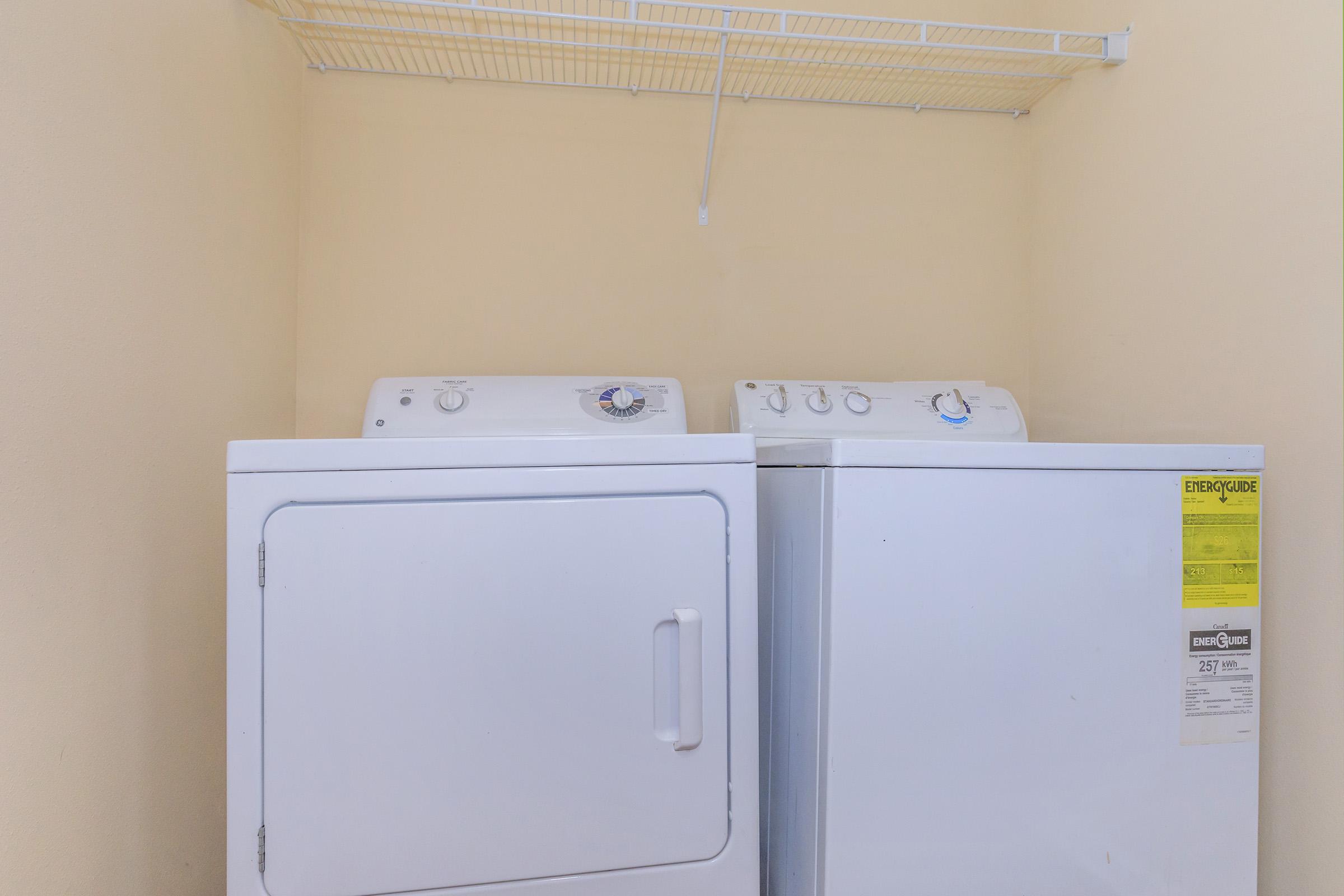 A stacked washer and dryer set located against a light-colored wall, with a wire shelf above for storage. The appliances are white and feature control dials on the front.