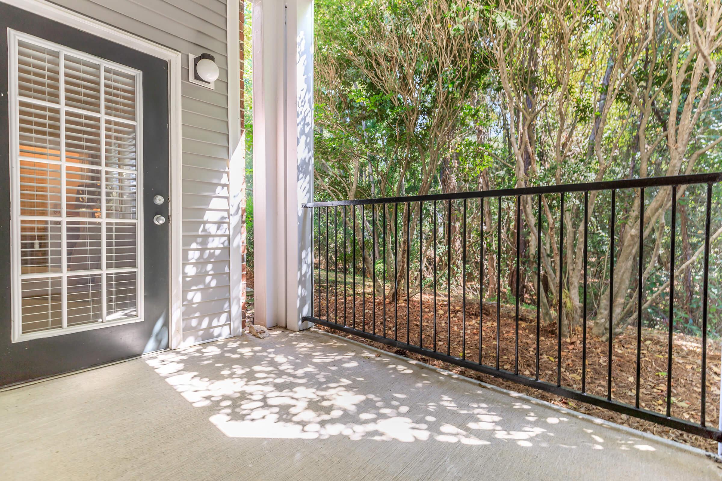 A small outdoor balcony or porch area featuring a black metal railing, light gray concrete flooring, and a door with glass panels. Surrounding the space are lush green bushes, casting dappled shadows on the ground from the sunlight.