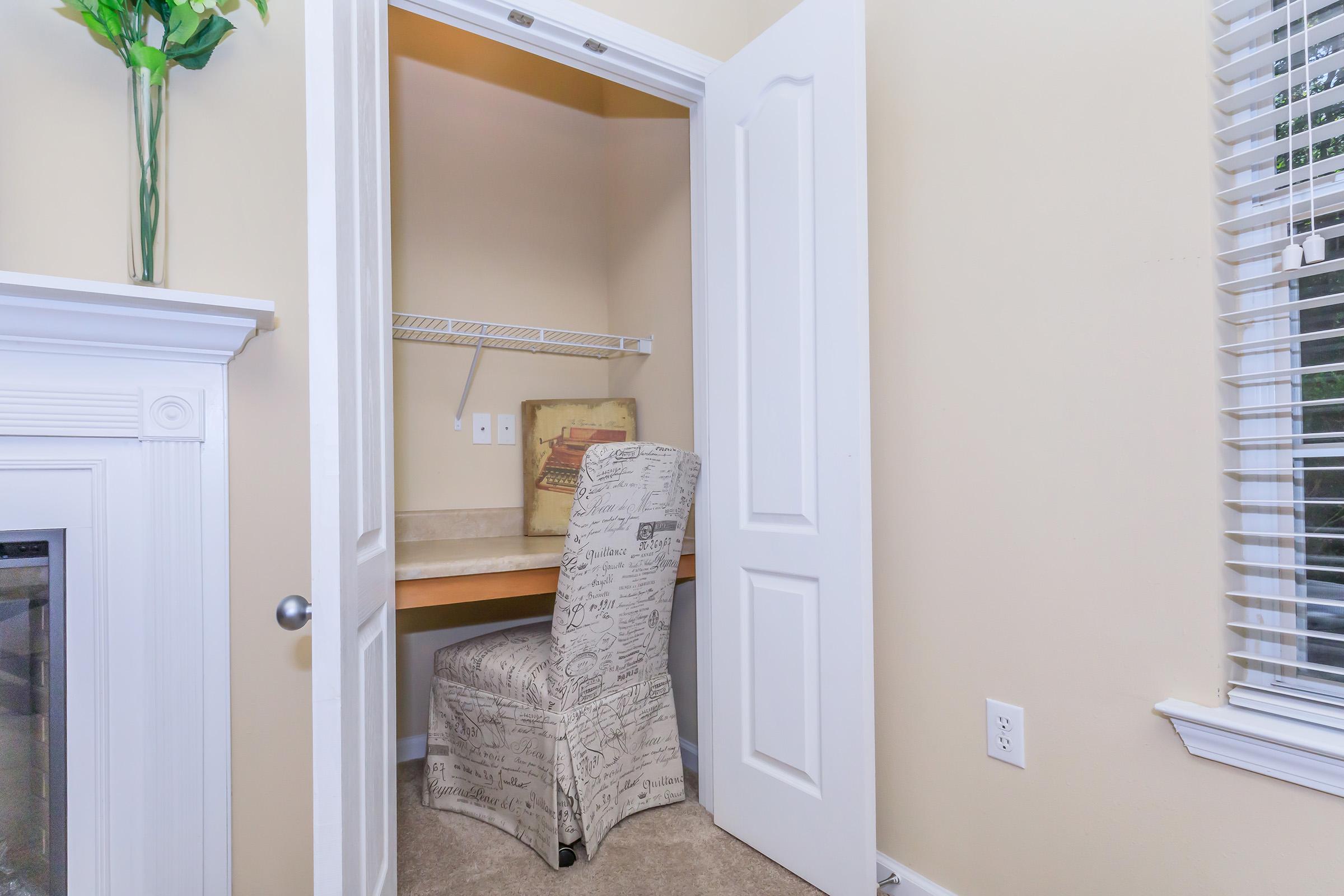 A small, well-lit nook with a beige wall, featuring an open closet door. Inside, there’s a wooden desk with a chair upholstered in a light fabric with printed text. Natural light streams in through nearby window blinds. Decorative flowers are placed on a mantle outside the nook.