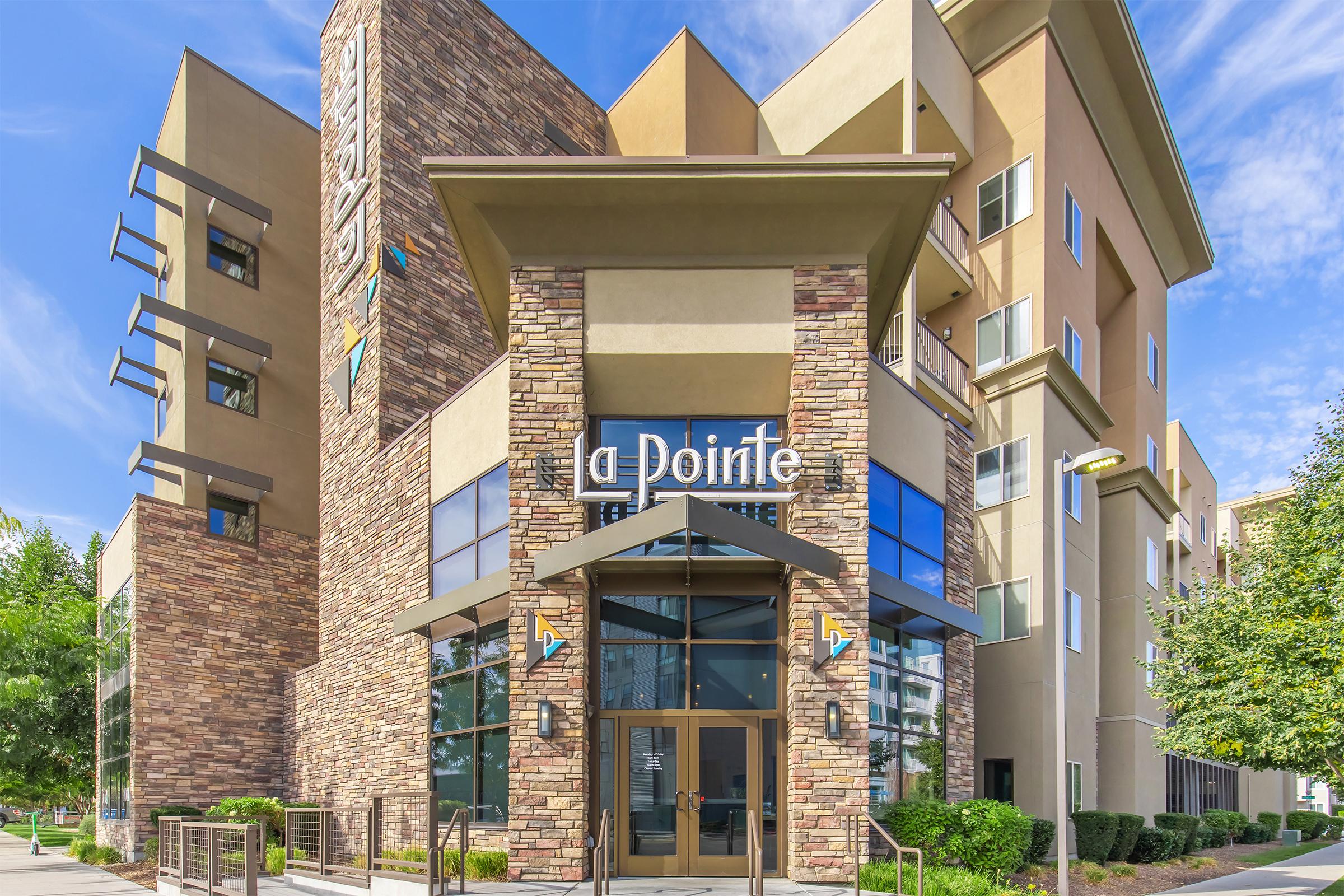 Modern apartment building with a stone and glass façade. The entrance features large glass doors and a sign that reads "a pointe." Surrounding landscaping includes green trees and shrubs, against a backdrop of bright blue skies.