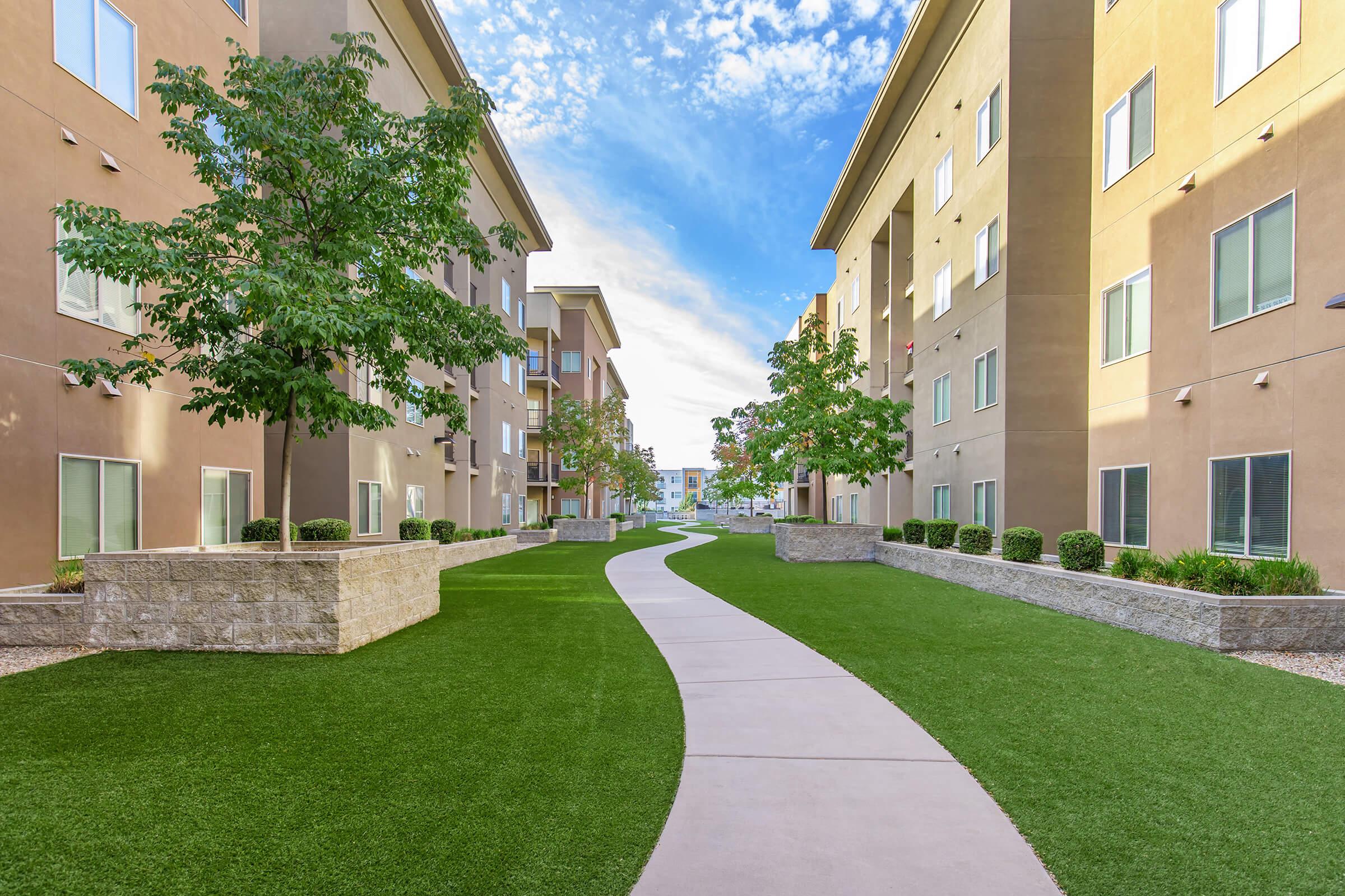 A well-maintained walkway lined by green grass stretching between two modern apartment buildings. Trees and shrubs are planted along the path, providing a pleasant, inviting atmosphere. The sky is partly cloudy, highlighting a bright and clear day.