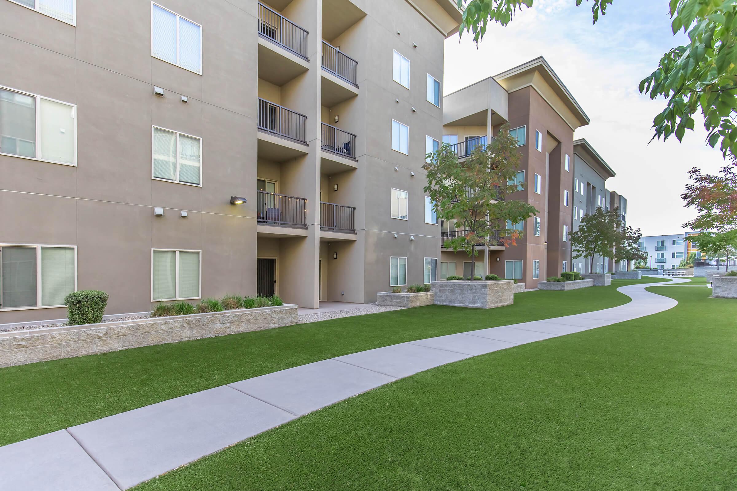 A view of a modern apartment complex featuring multiple buildings with balconies, surrounded by well-manicured green lawns and a smooth sidewalk. The scene is bright, showcasing a clear sky and landscaped greenery, indicating a peaceful residential environment.