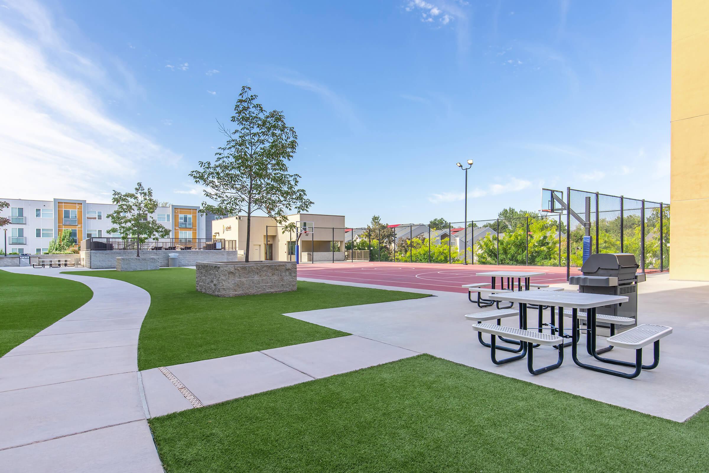 A modern outdoor space featuring a basketball court, picnic tables, and green landscaping. There are walkways leading through the area, and nearby buildings are visible in the background. The setting appears inviting and well-maintained for recreational activities.