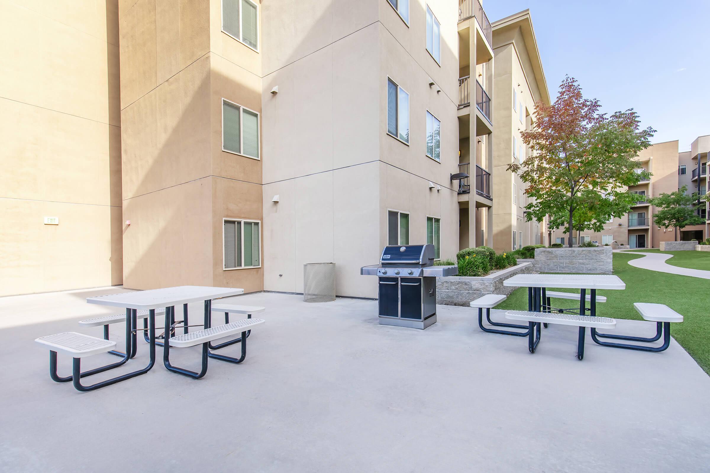 Outdoor BBQ area featuring a grill and picnic tables, set against a backdrop of beige apartment buildings and greenery. The space is well-lit with open skies, providing a communal area for residents to gather and enjoy meals outside.