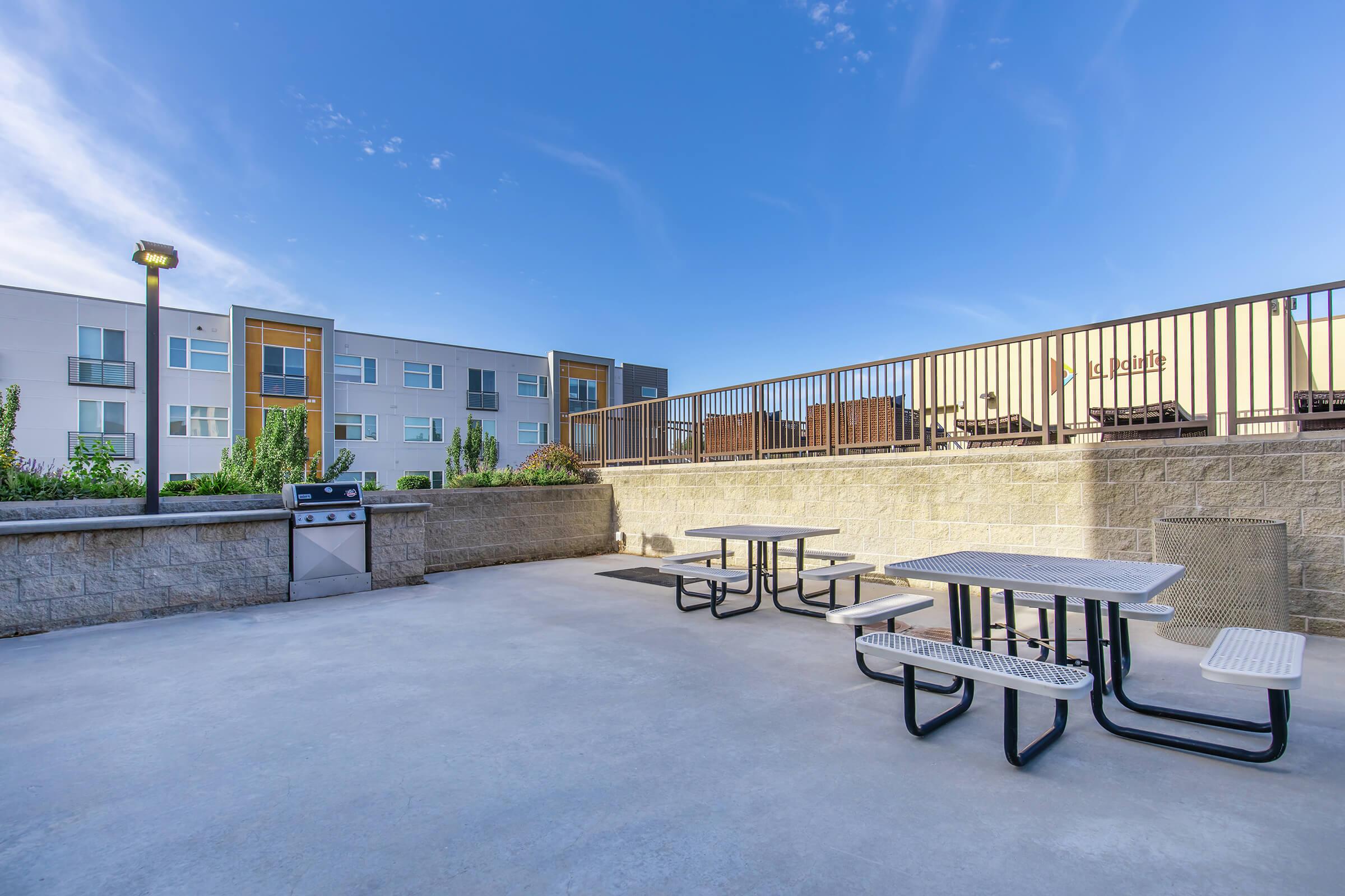 Outdoor patio area featuring multiple picnic tables and a barbecue grill, surrounded by low landscaping and with an apartment building in the background under a clear blue sky. The space is designed for relaxation and social gatherings.