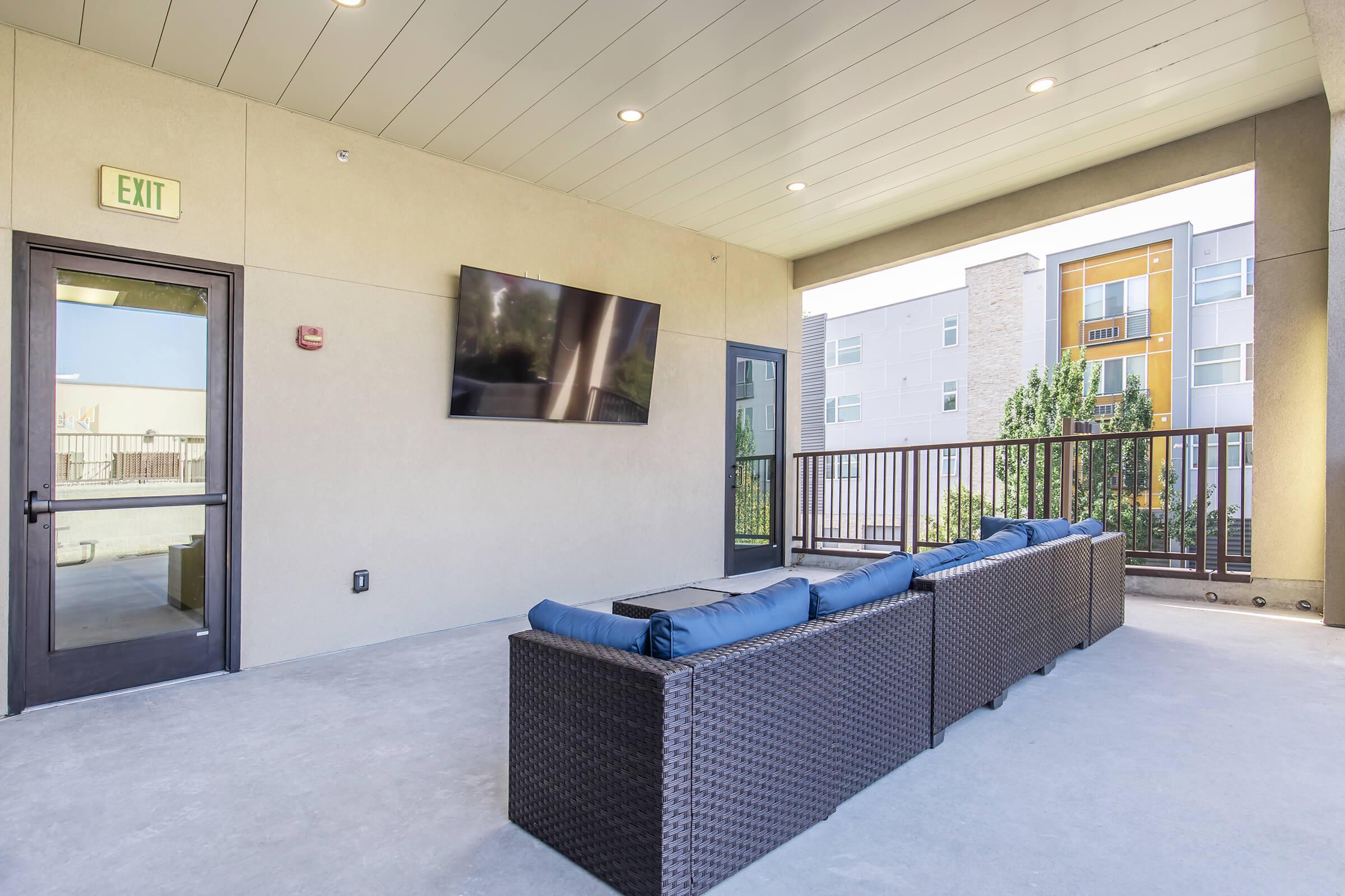A modern outdoor lounge area with a dark wicker sofa featuring blue cushions, facing a wall-mounted television. The space is covered and includes a glass door leading to another area, with greenery visible in the background alongside multi-story residential buildings.