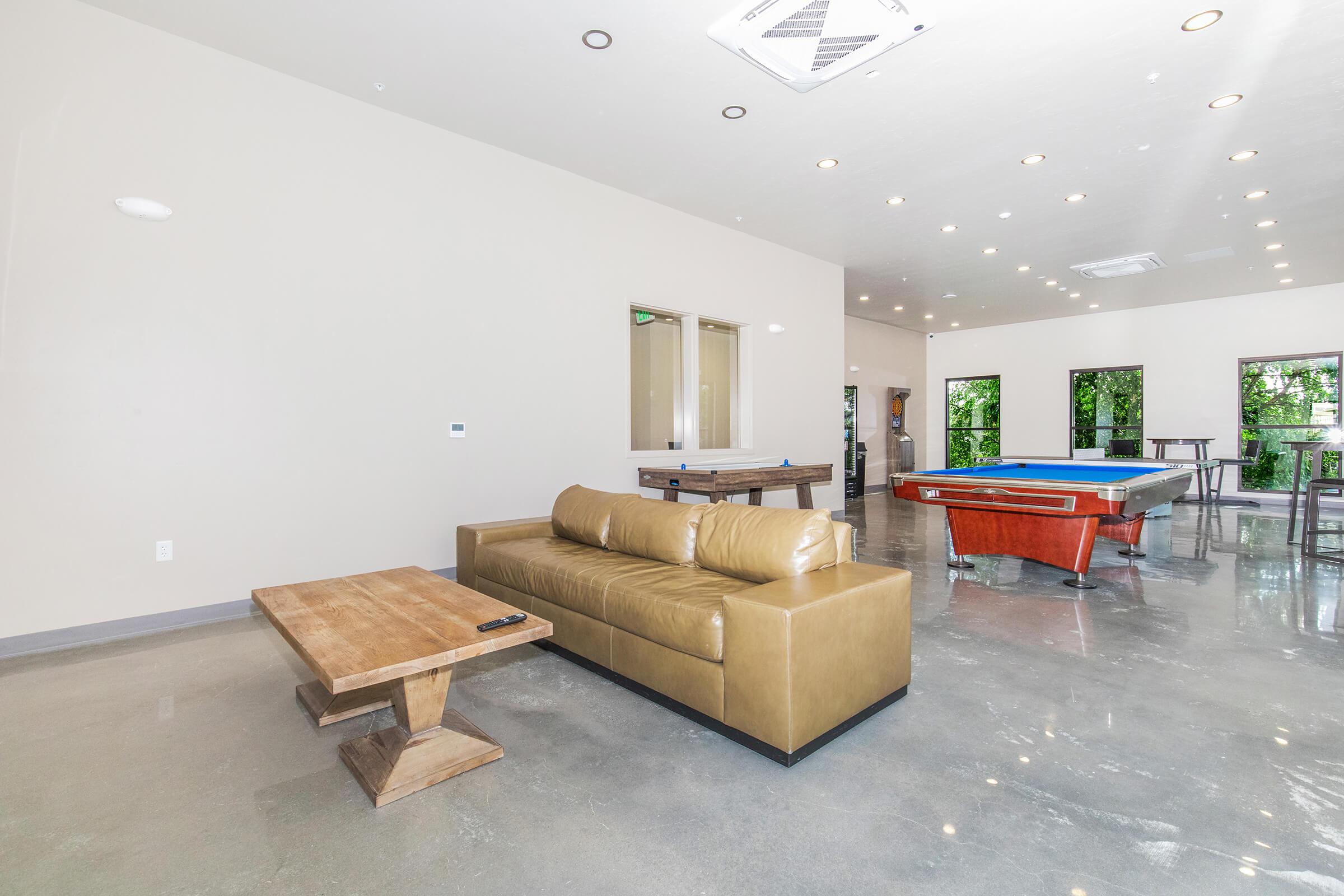 A spacious interior with a light-colored wall and polished concrete floor. A modern brown leather sofa is placed in front of a wooden coffee table. In the background, there are large windows allowing natural light and a blue pool table near a bar-style seating area.