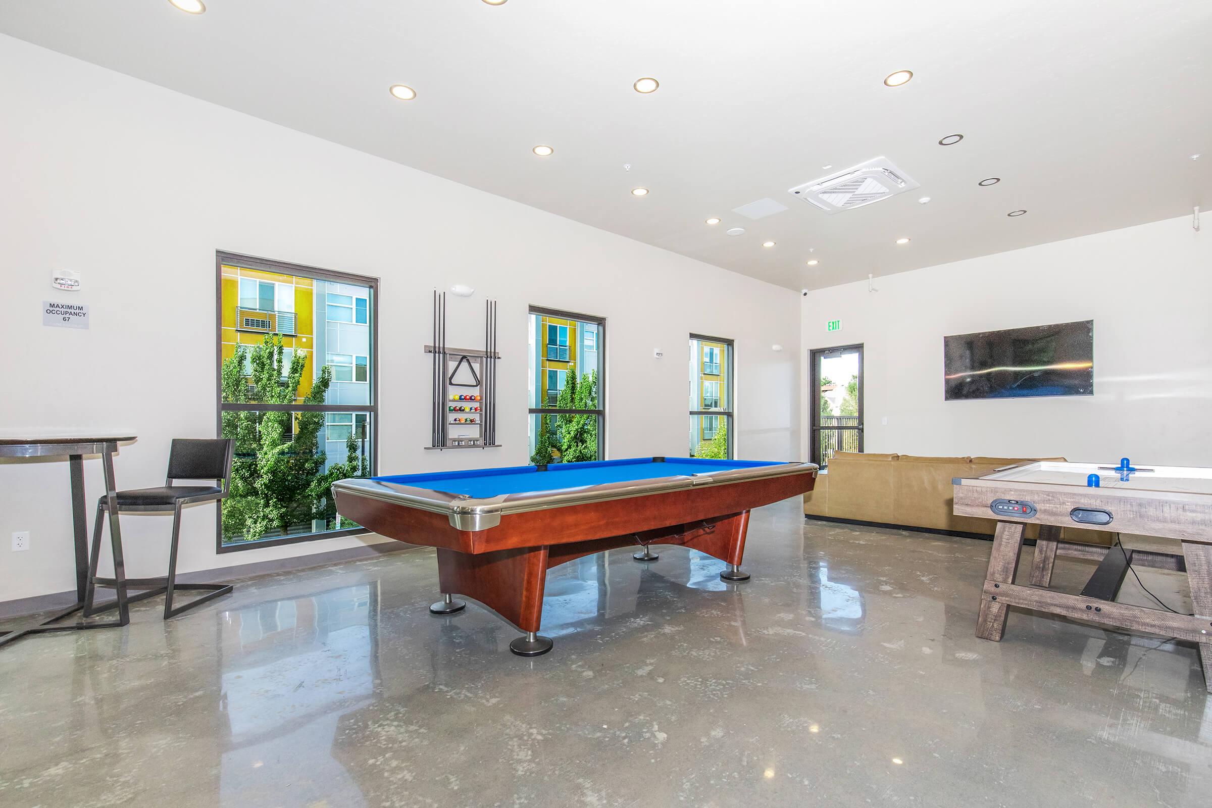 A modern game room featuring a blue felt pool table with wooden legs, a sophisticated beige sofa, and a foosball table. Large windows allow natural light to fill the space, showcasing greenery outside. The polished concrete floor adds to the contemporary aesthetic.