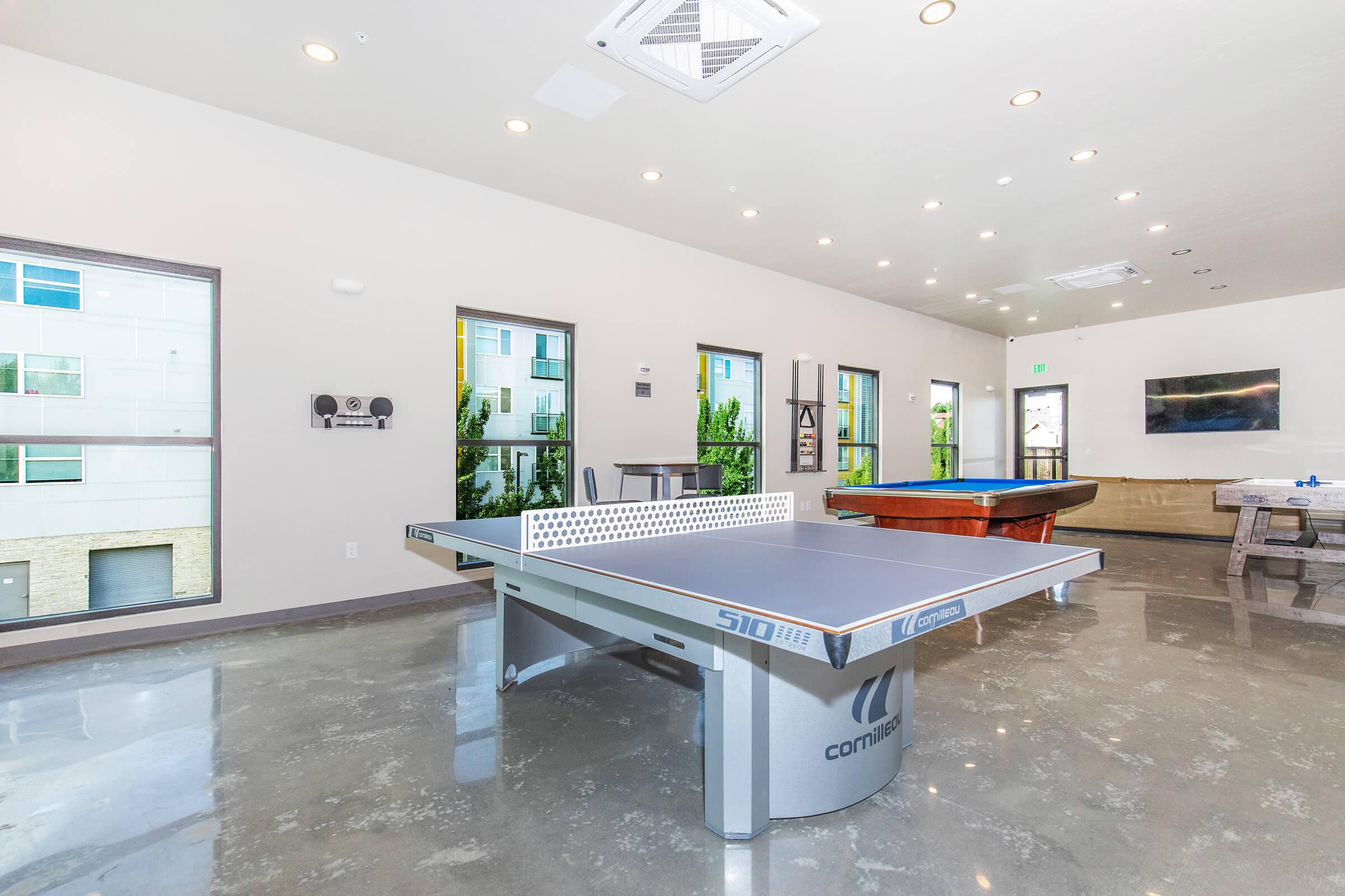 A spacious recreational room featuring a table tennis table in the center, surrounded by large windows letting in natural light. There are various games including a pool table and a seating area, with modern decor and polished concrete floors.