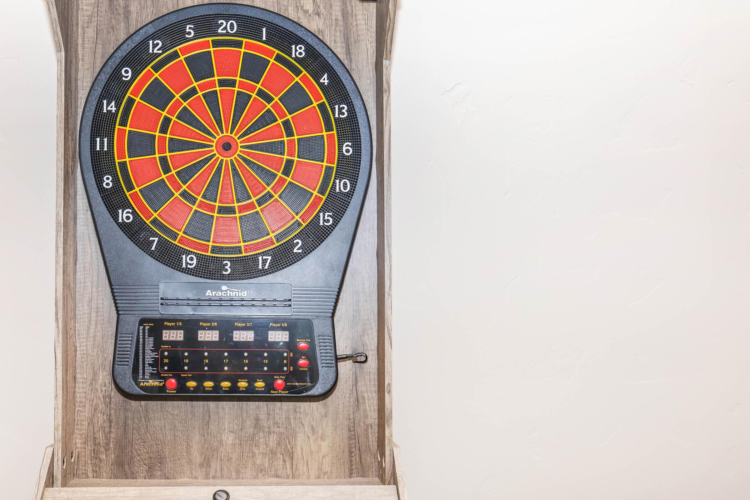 Electronic dartboard mounted on a wooden wall, featuring a colorful target with red, green, and yellow sections. Below the target are digital score displays and control buttons for gameplay. The background is a simple, light-colored wall.