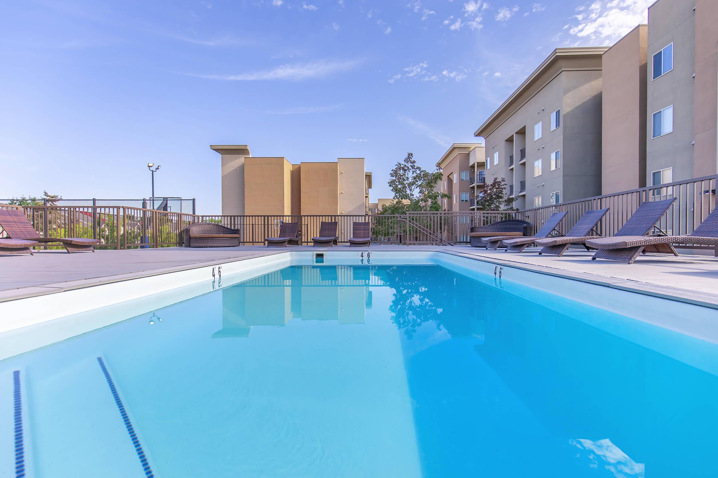 A clear blue swimming pool surrounded by lounge chairs, with a modern multi-story building in the background. The sky is bright with a few clouds, creating a peaceful and inviting atmosphere for relaxation and leisure.