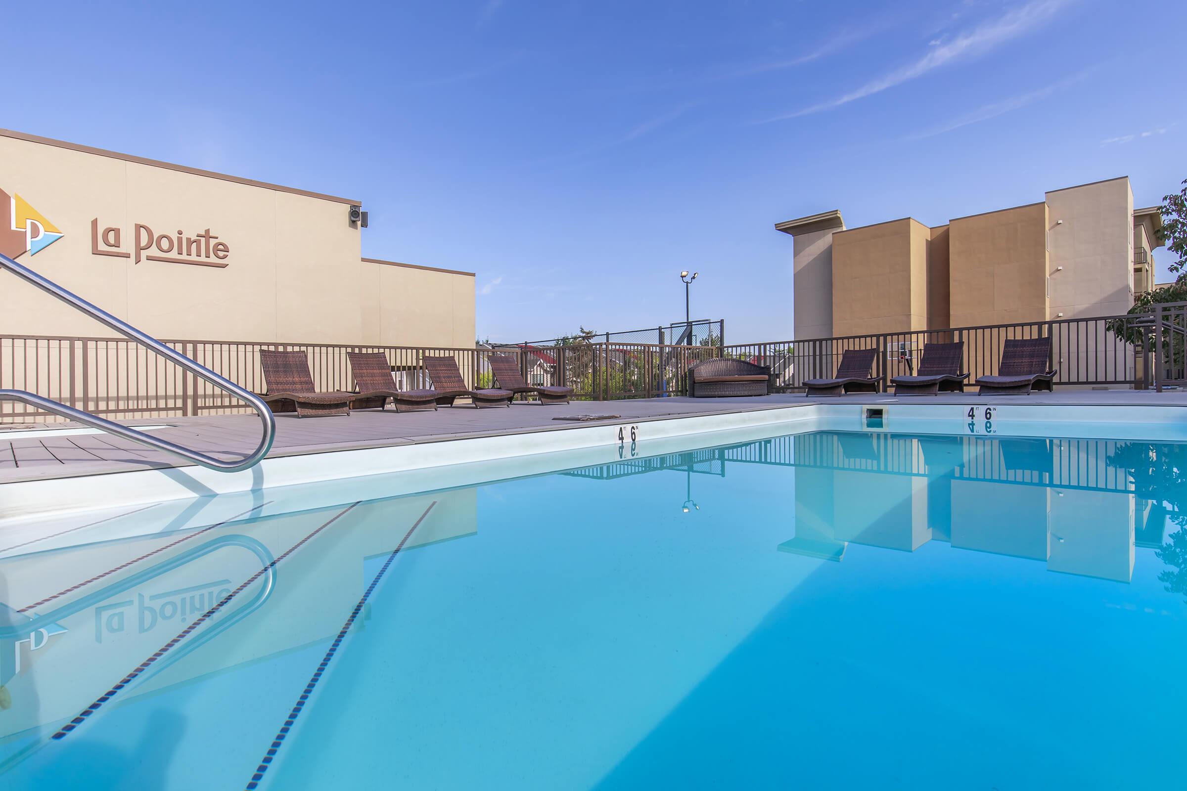 A rooftop pool area with clear blue water reflecting the sky. Sun loungers are placed around the pool's edge, and a building labeled "La Pointe" is visible in the background. The scene is bright and inviting, perfect for relaxation. The sky is mostly clear with a few clouds.