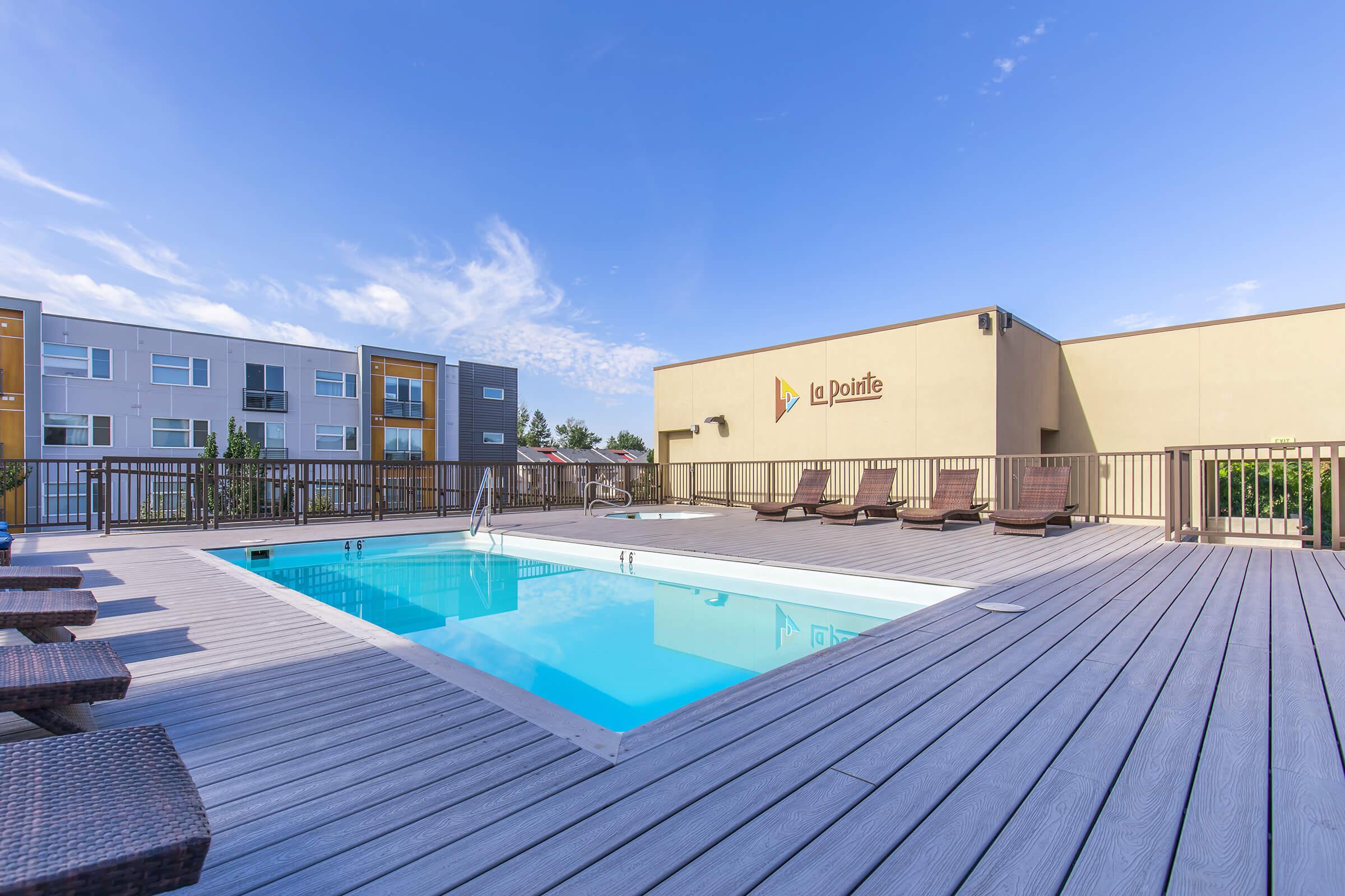 View of a residential pool area featuring a clean, blue swimming pool surrounded by wooden decking. Several lounge chairs are positioned around the pool, with modern apartment buildings in the background under a clear blue sky. The sign "Le Pointe" is visible on the building wall.