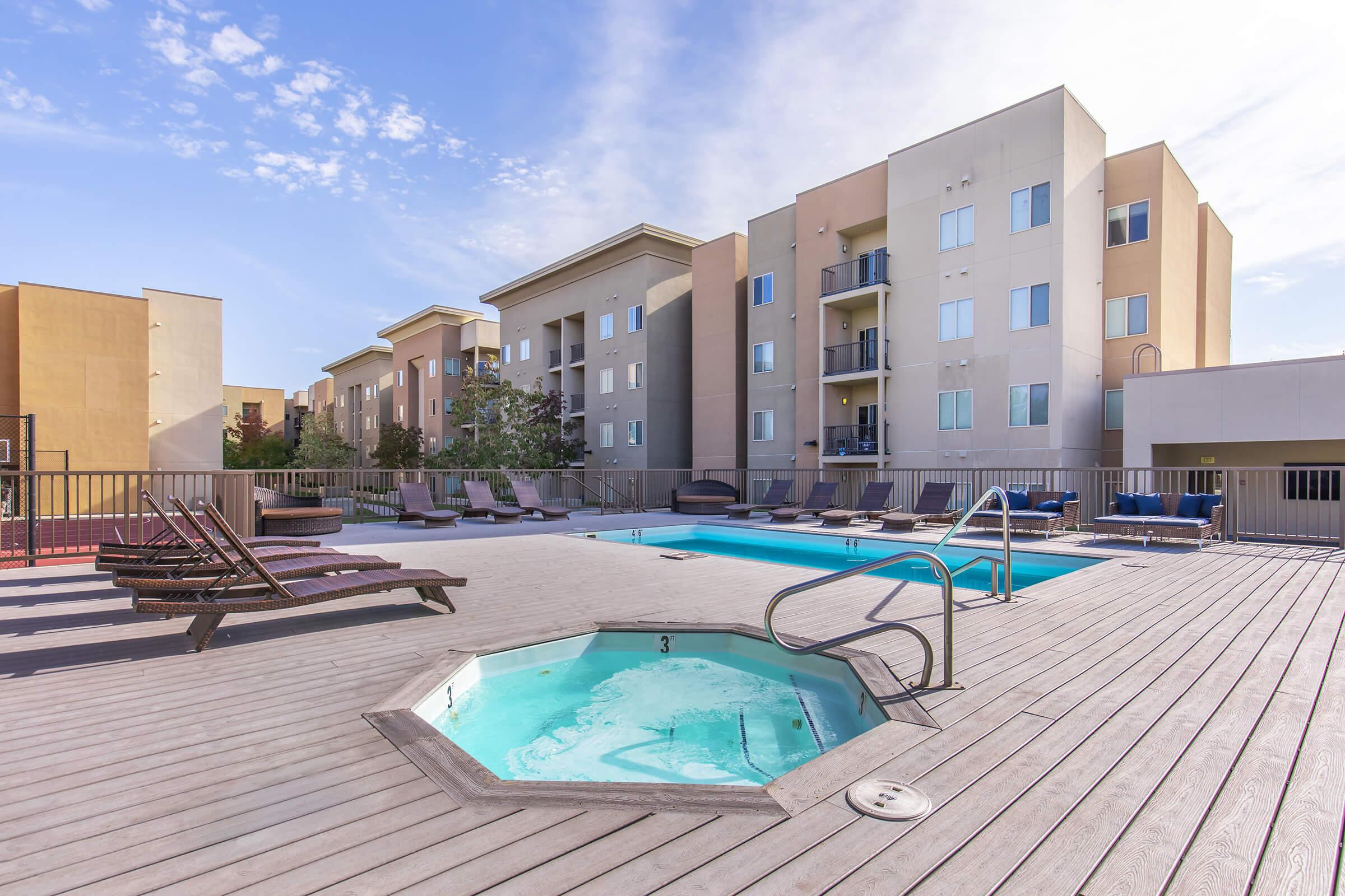 A modern outdoor swimming pool area featuring an octagonal hot tub, surrounded by wooden decking and lounge chairs. The backdrop includes beige apartment buildings with balconies, under a clear blue sky with scattered clouds.
