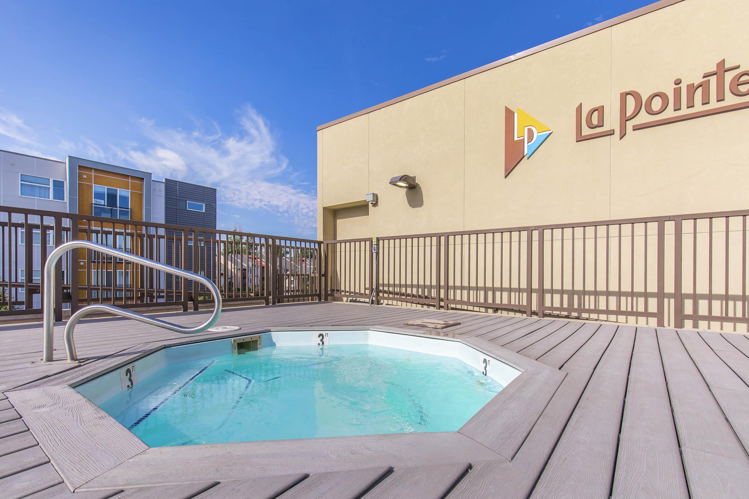 Hot tub on a rooftop deck, surrounded by a wooden railing. The background features modern buildings with colorful accents under a clear blue sky. The hot tub area is well-lit, creating a welcoming atmosphere for relaxation.