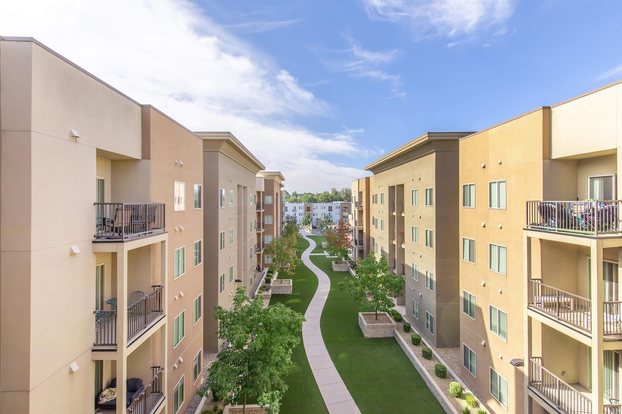 Aerial view of a modern apartment complex with beige and tan buildings flanking a landscaped pathway. Lush green grass and trees line the walking path, which leads through the development. The sky is partly cloudy, adding to the serene atmosphere of the community setting.
