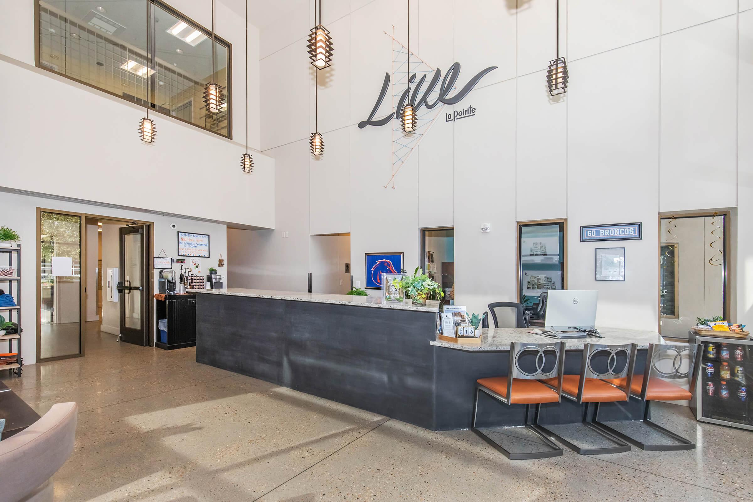 A modern reception area with a sleek front desk, featuring a sign that reads "Line" above it. The space has high ceilings, contemporary lighting fixtures, and large windows. There is a computer on the desk, plants for decoration, and seating areas with chairs visible in the foreground.