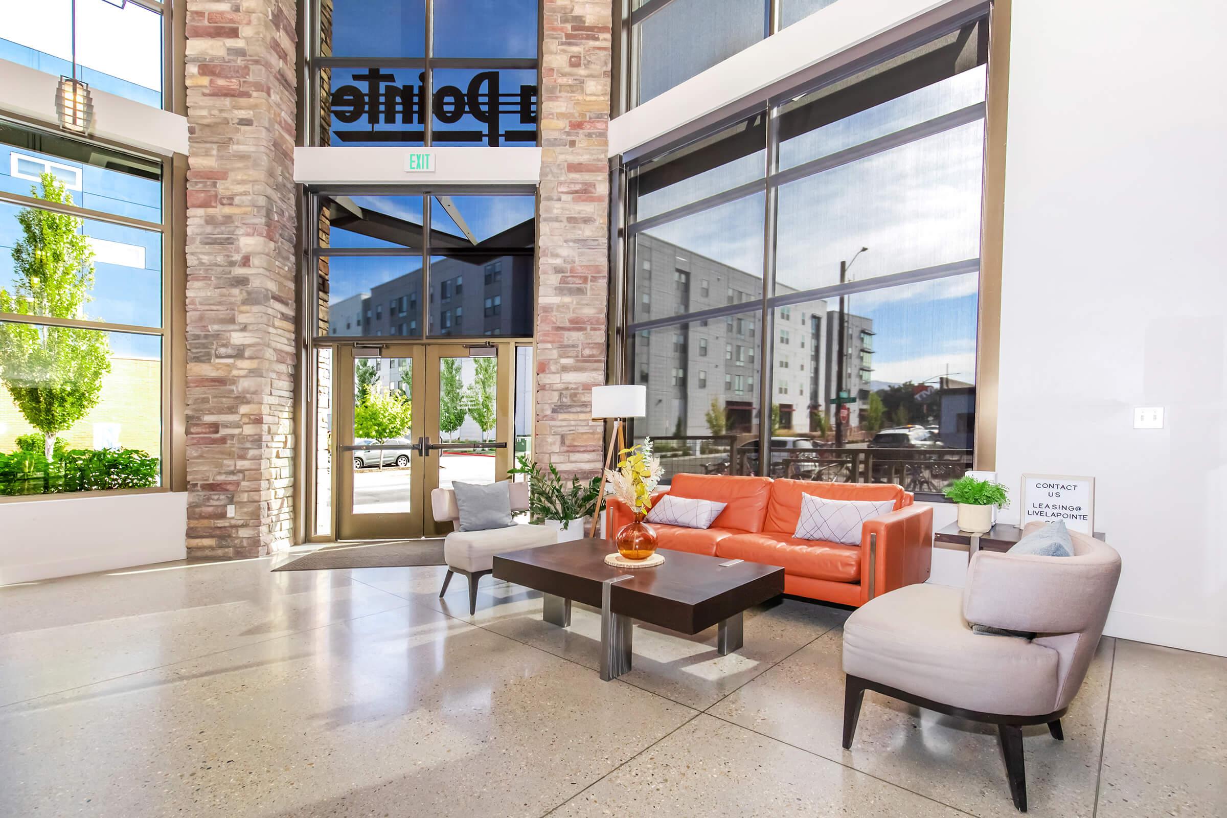 Modern lobby area featuring large windows, stone accents, and a mix of seating. An orange sofa, a coffee table with decorative flowers, and two stylish chairs create a welcoming atmosphere. Natural light brightens the space, showcasing greenery outside.