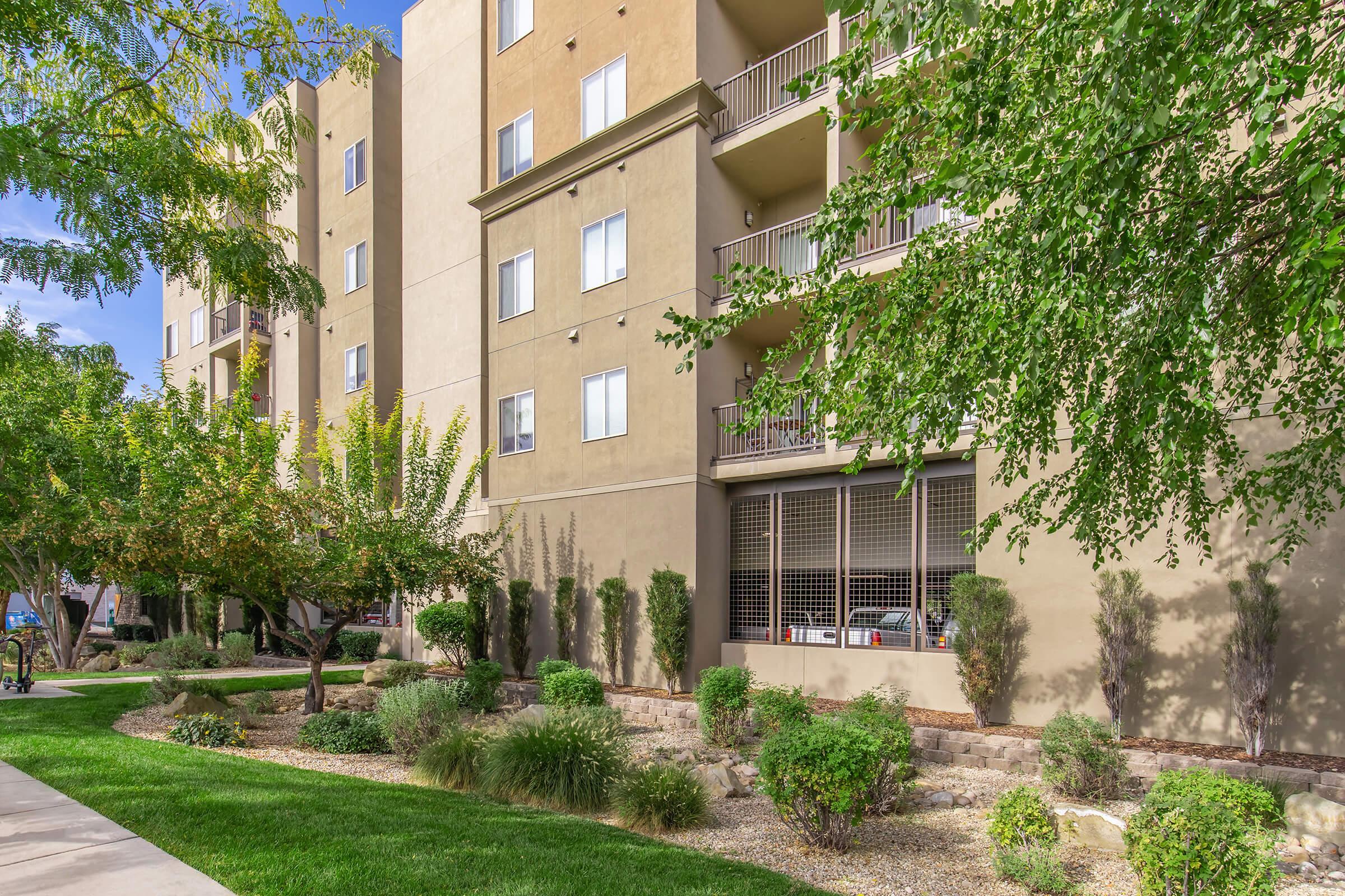 A multi-story apartment building with a beige exterior, featuring balconies and large windows. Lush green landscaping with various shrubs and small trees surrounds the building, and a paved walkway leads through the grass. The sky is clear, suggesting a sunny day.