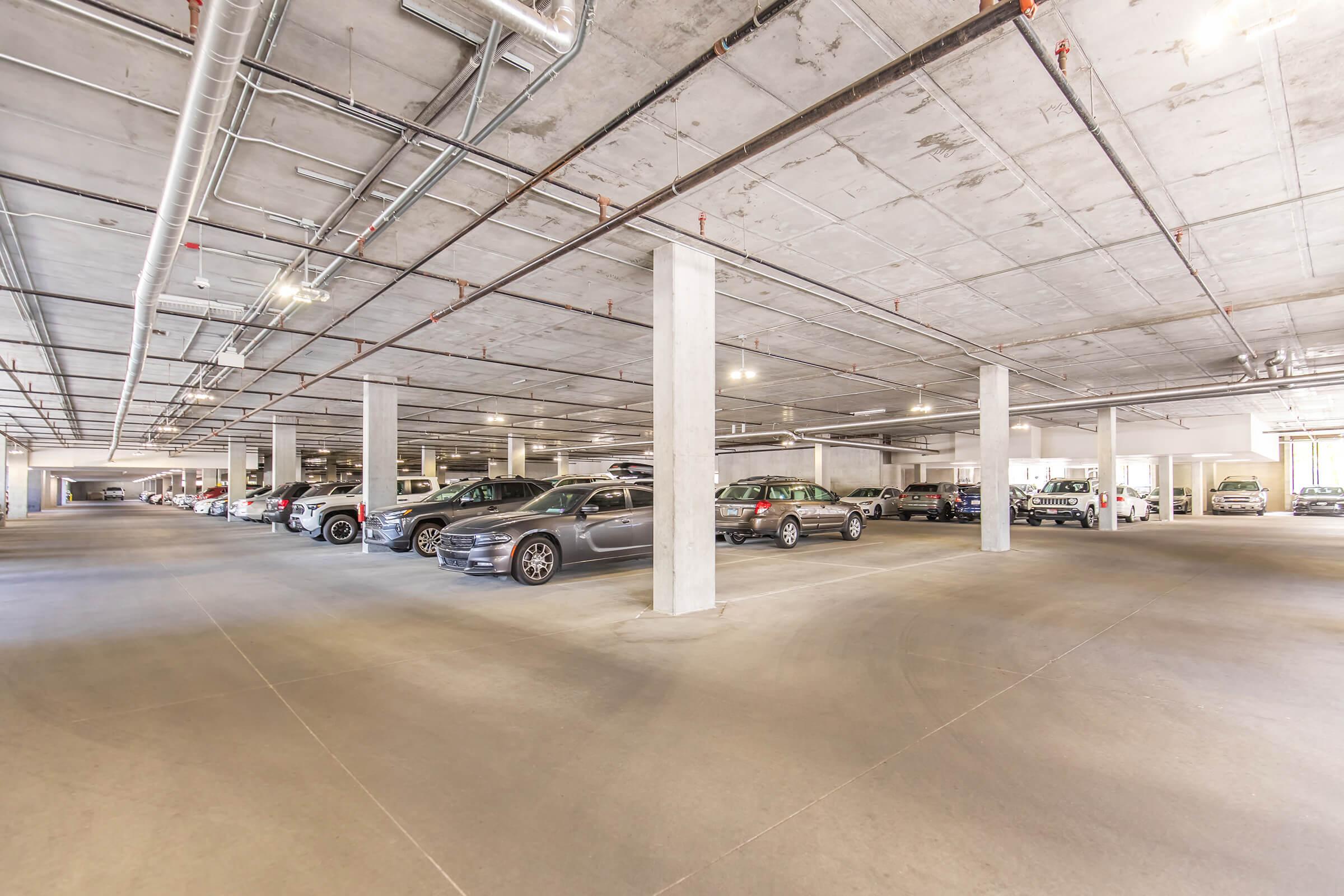 A spacious underground parking garage featuring multiple rows of parked cars. The area has concrete floors, exposed pipes, and bright overhead lighting. Several concrete columns are positioned throughout the space, providing structural support. There are clear driving lanes and empty parking spots visible.