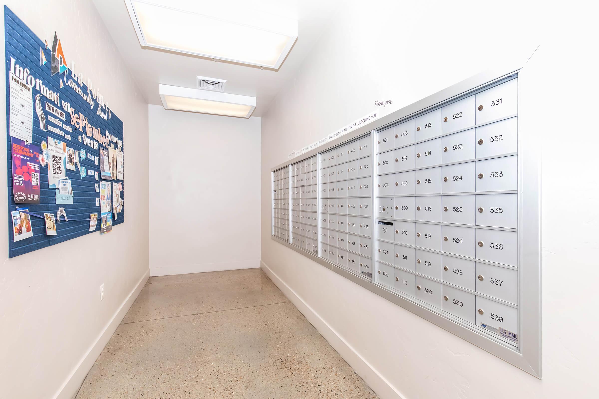 A hallway featuring a bulletin board with information and announcements on the left and a row of metallic mailboxes on the right, numbered from 531 to 555. The space is well-lit with a bright overhead light, and the walls and floor have a simple, clean design.